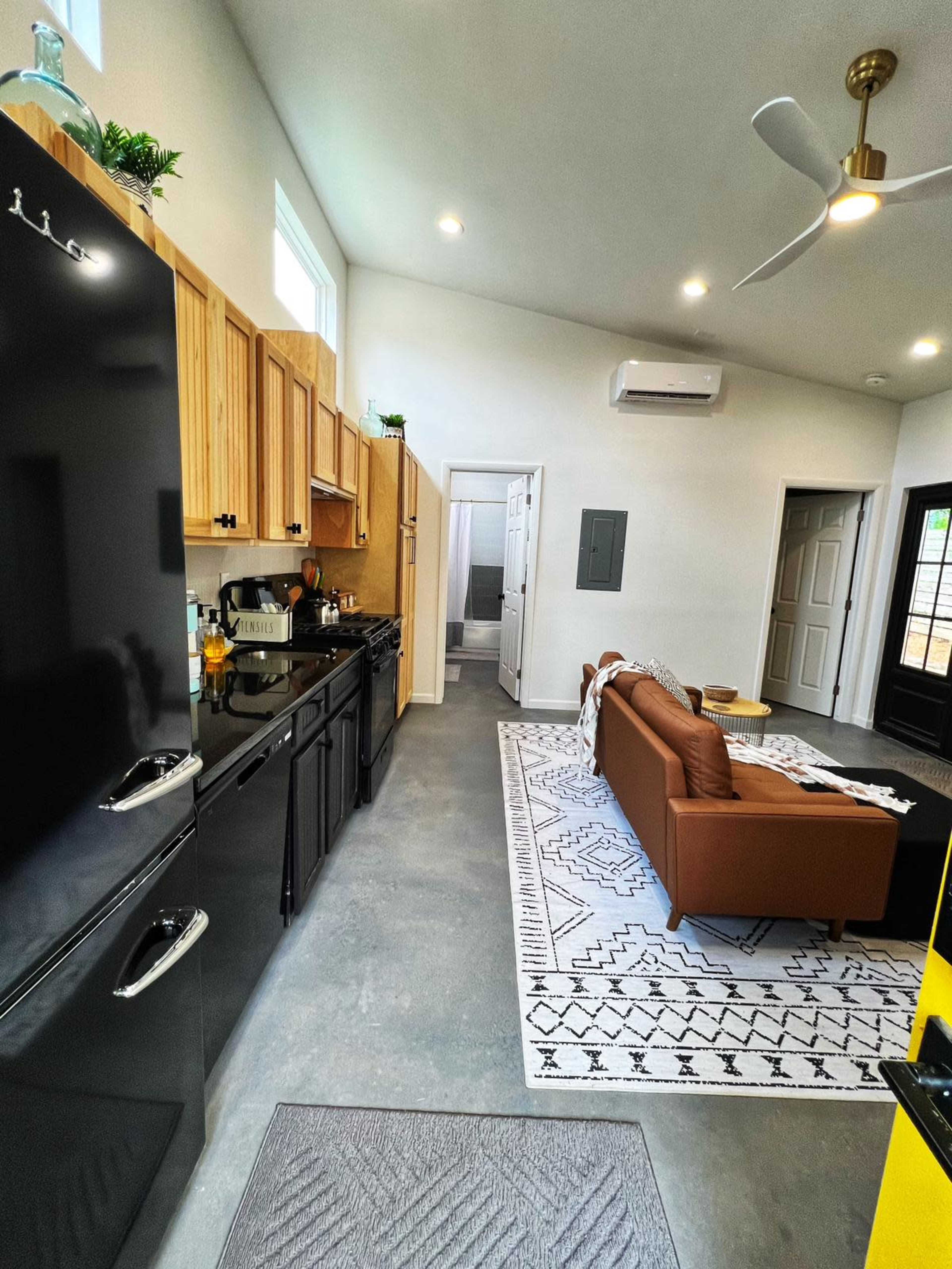 A modern kitchen and living area, featuring a black refrigerator, wooden cabinets, and a brown sofa on a patterned rug.