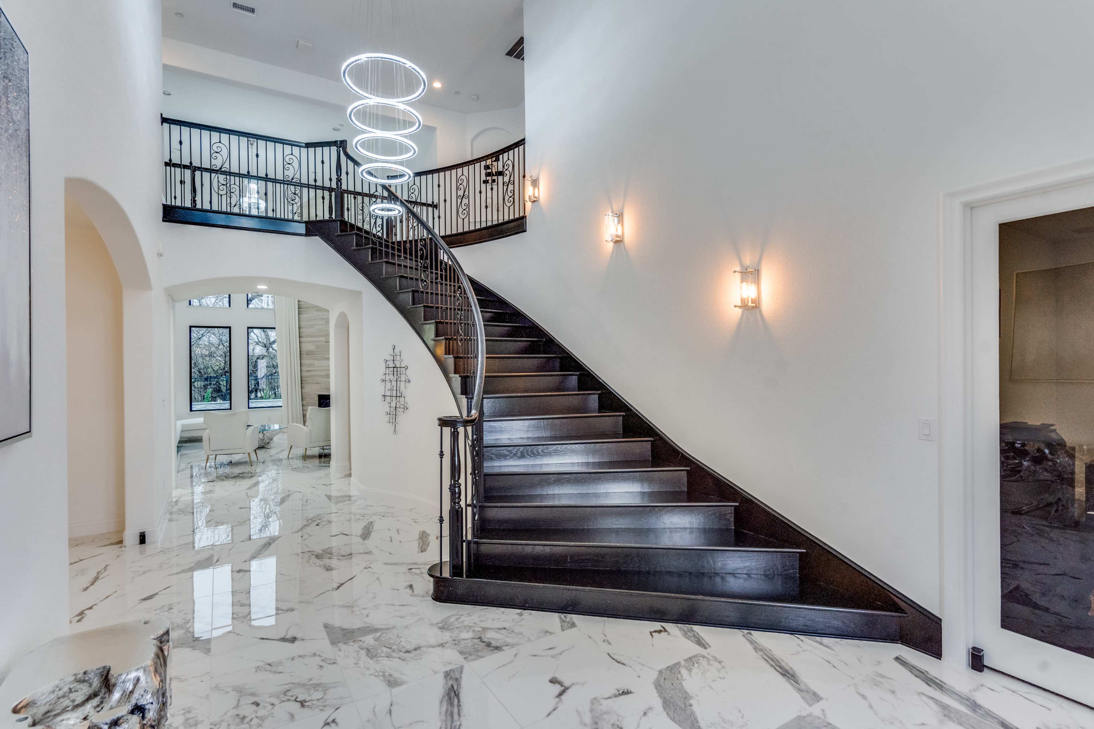 The image shows a spacious foyer featuring a curved wooden staircase with a modern chandelier above and glossy marble flooring.