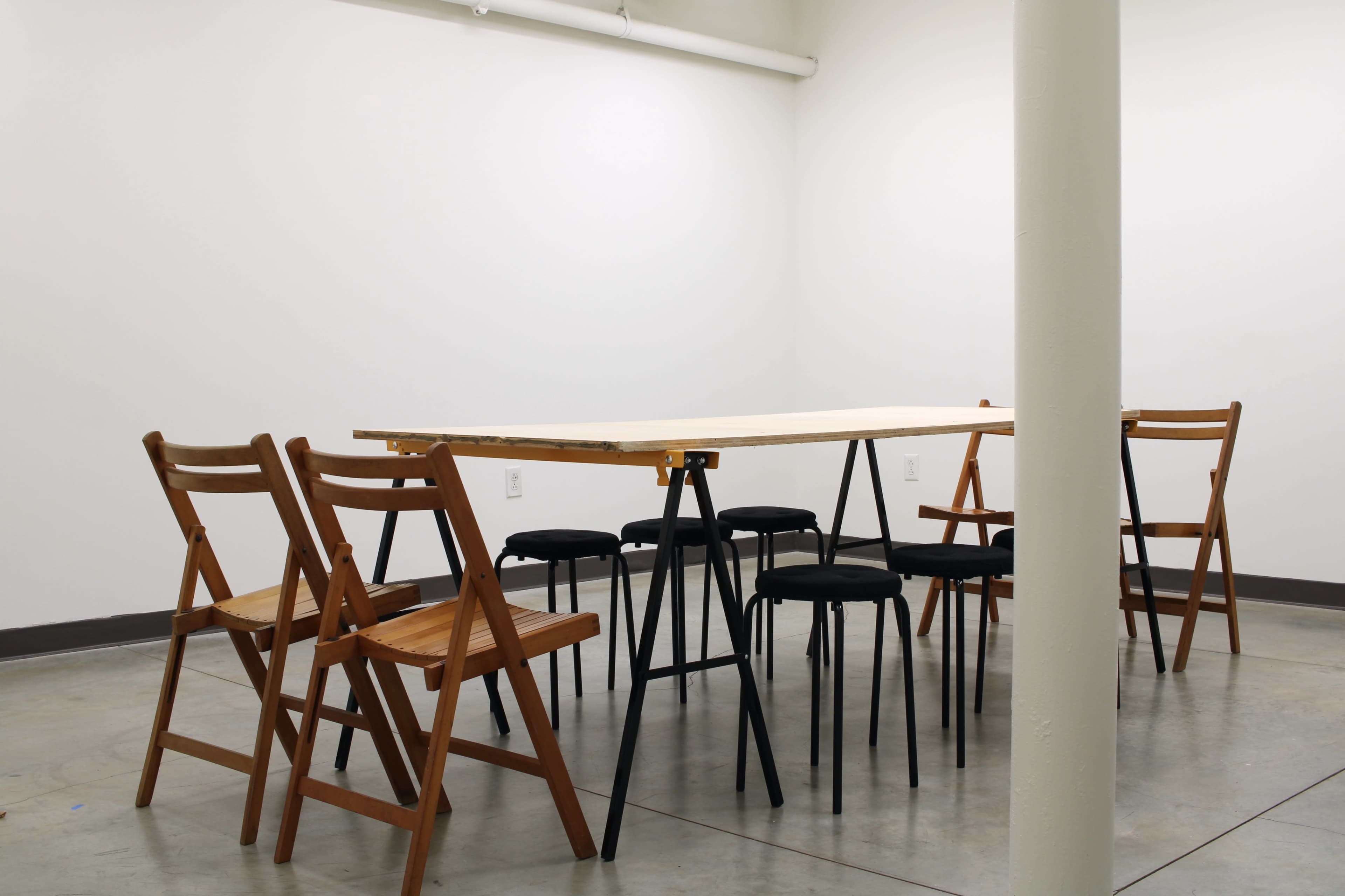 A wooden table is surrounded by six chairs in a sparsely decorated room with a concrete floor and white walls.