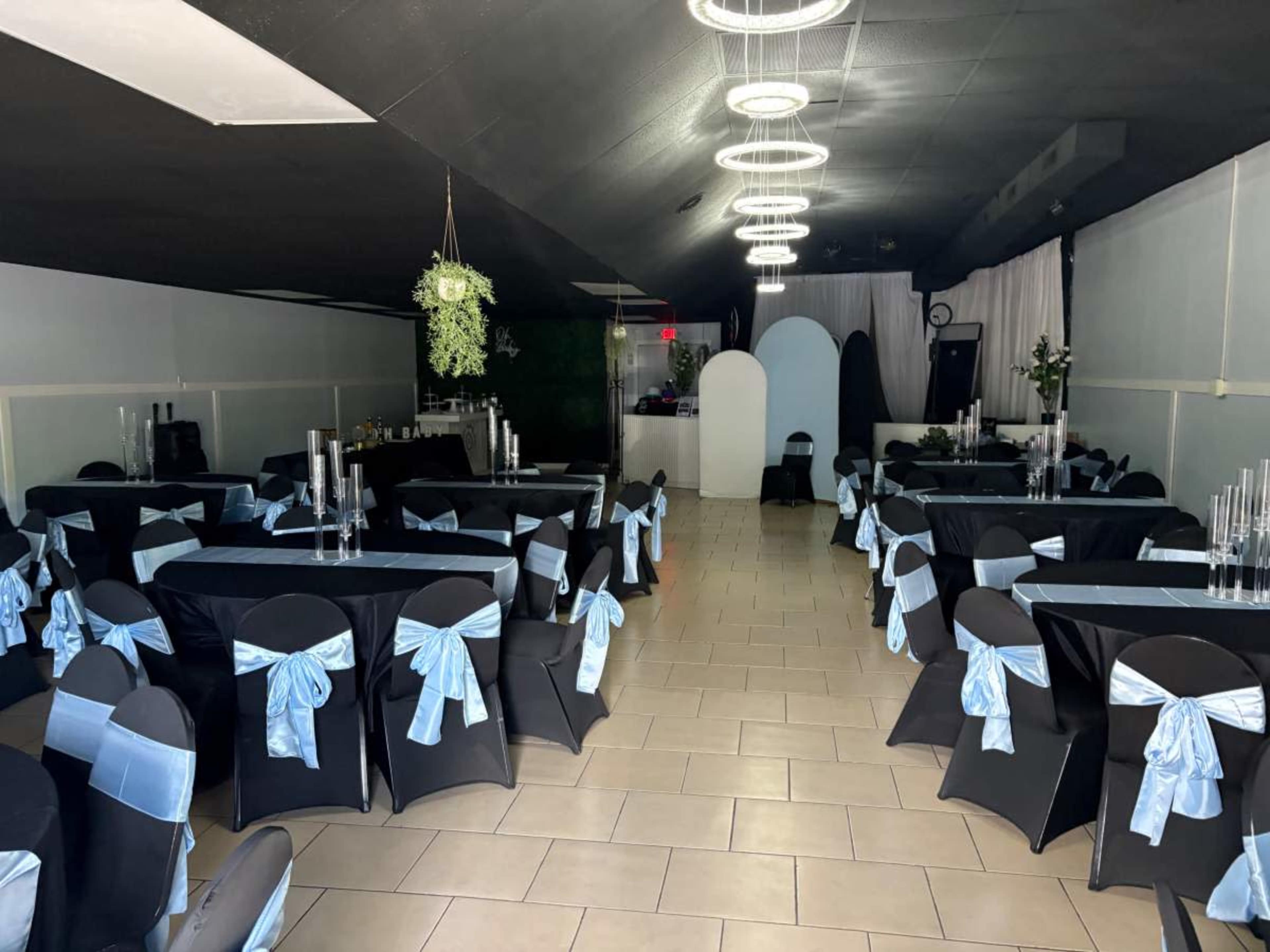 The image shows a large event space set up with black tables covered with light blue sashes and tall glass centerpieces, alongside a bar area in the background.