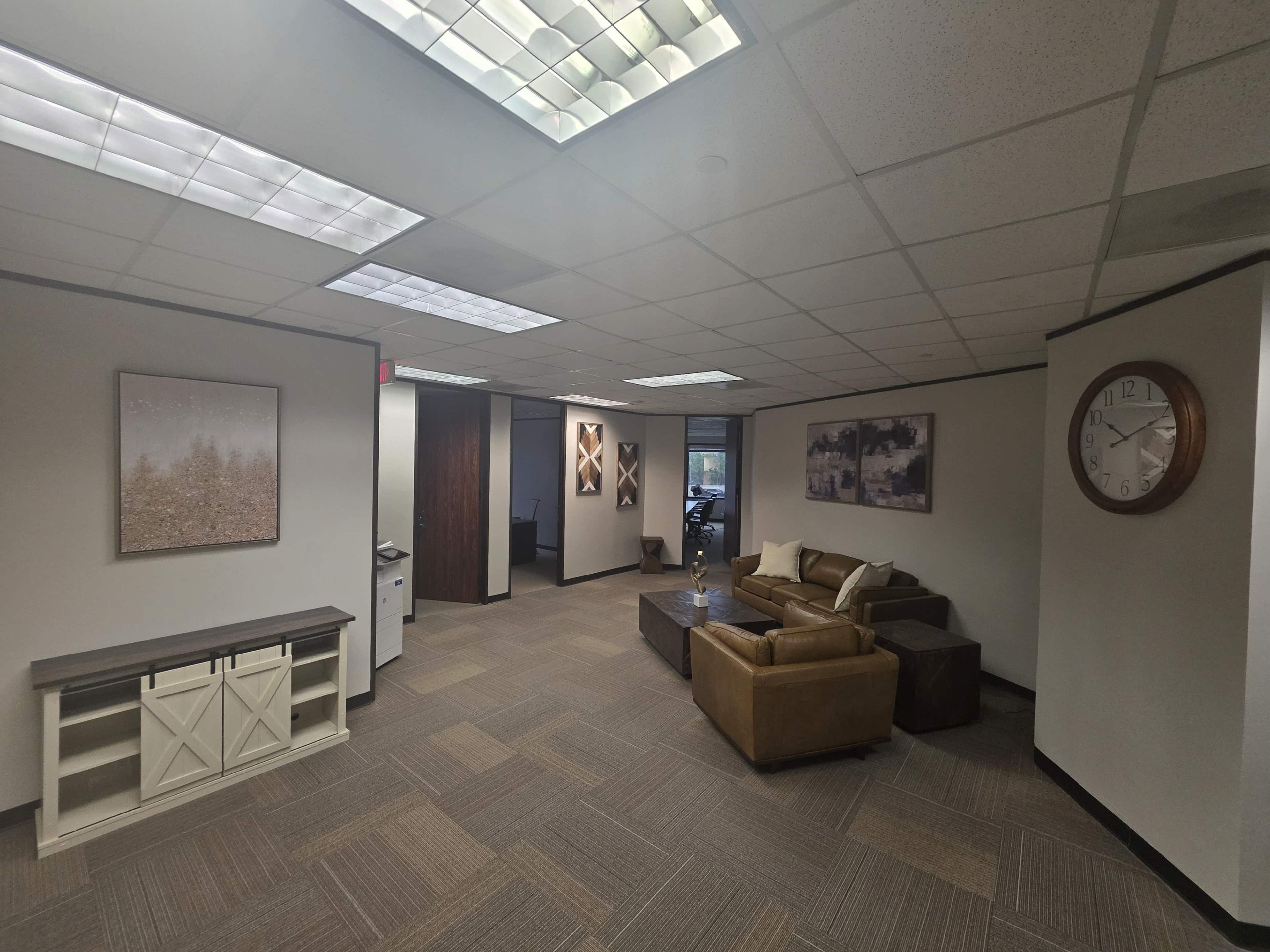 The image shows an office interior with a seating area, featuring a brown sofa, coffee table, and decorative wall art, along with a large clock and a reception area visible in the background.