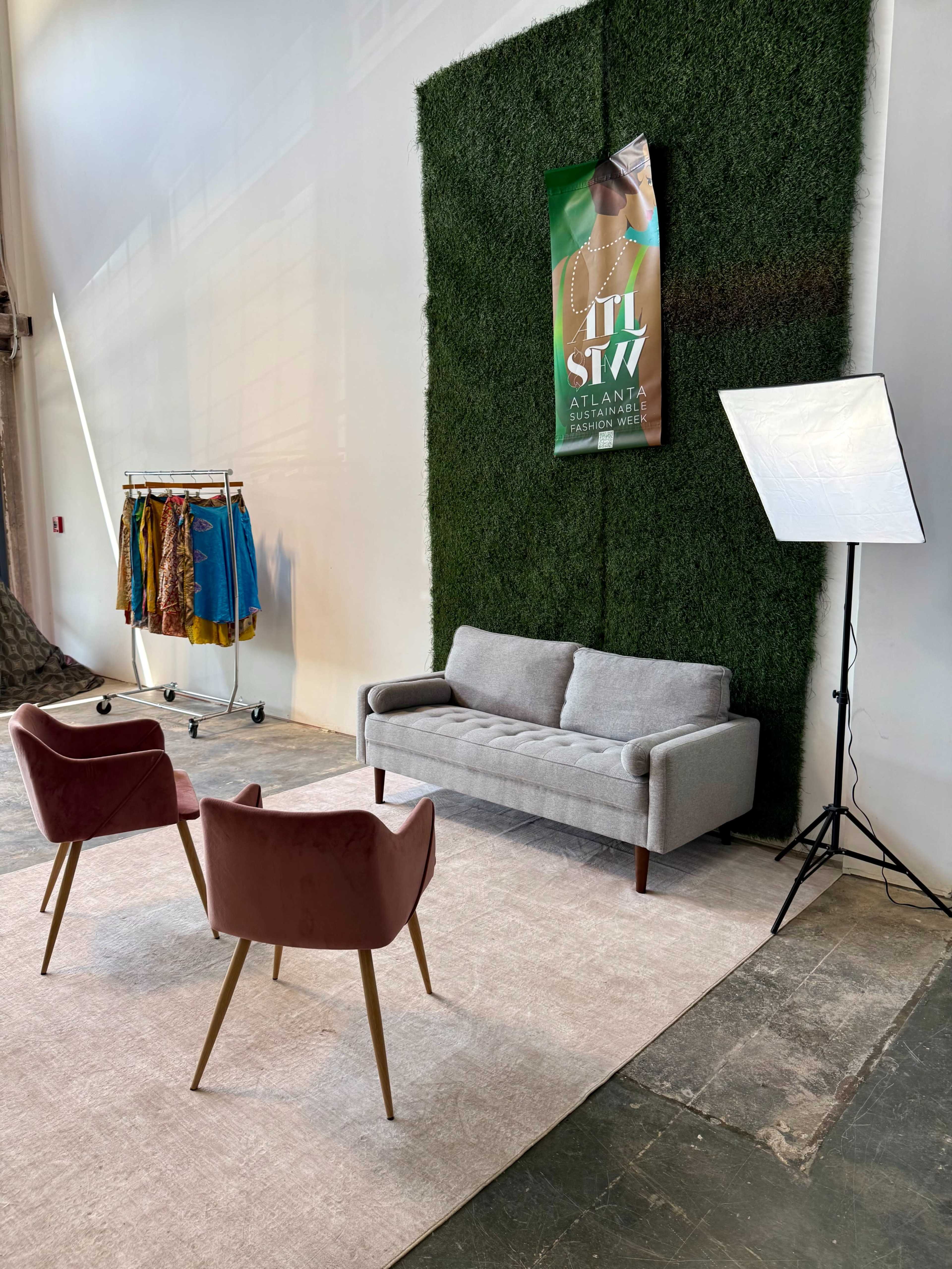 The image shows a minimalist setup featuring a gray sofa, two pink chairs, a clothing rack with colorful garments, and a backdrop of green foliage, alongside a light source.