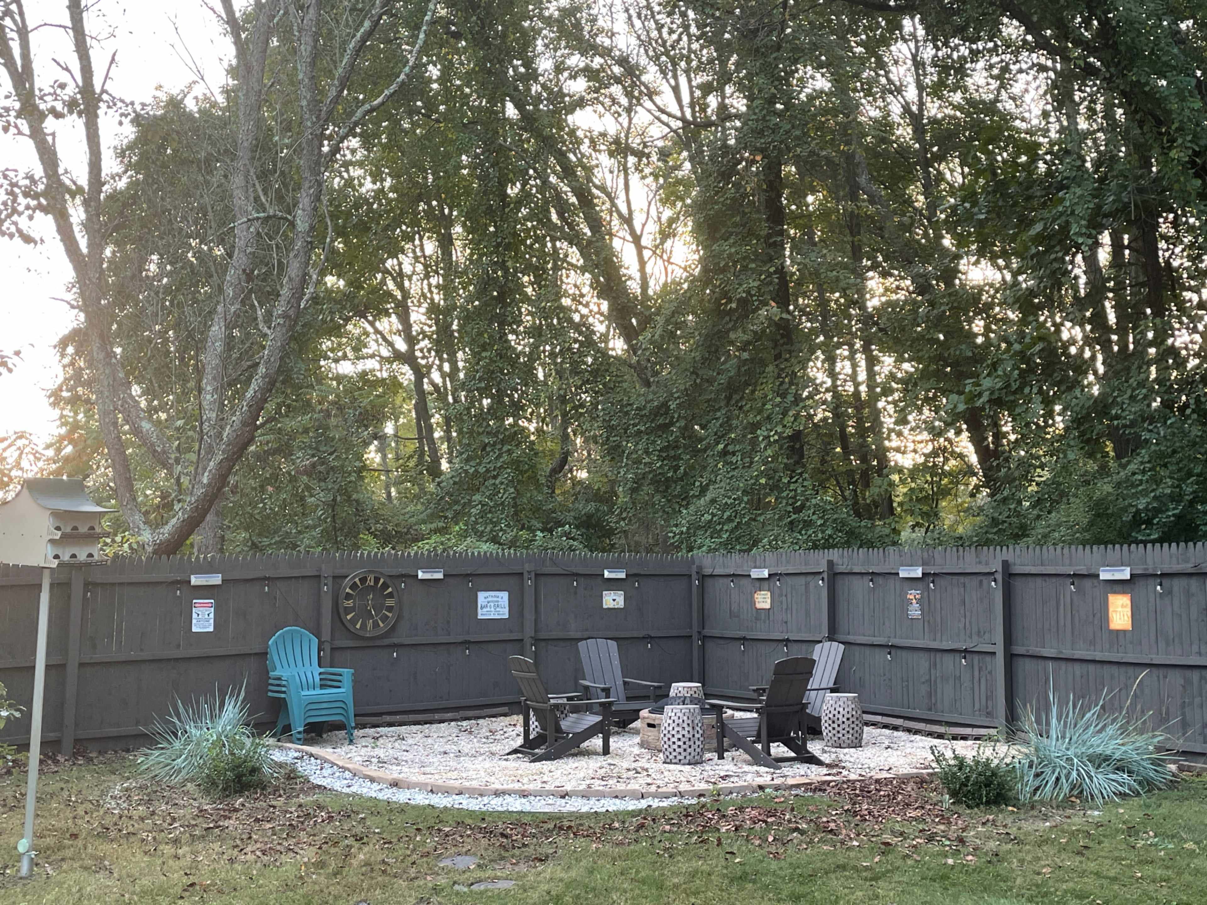The image shows a fenced outdoor area with seating, gravel, and decorative elements surrounded by trees.