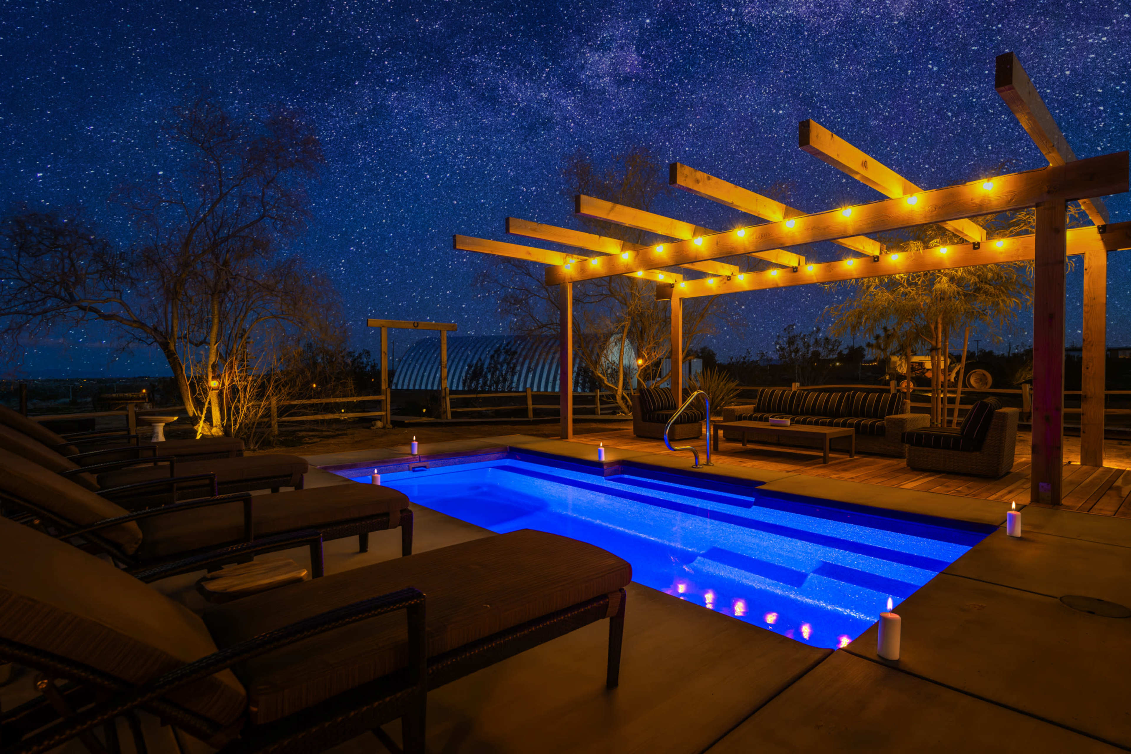 A modern backyard patio features a swimming pool illuminated by blue lights under a starry night sky.