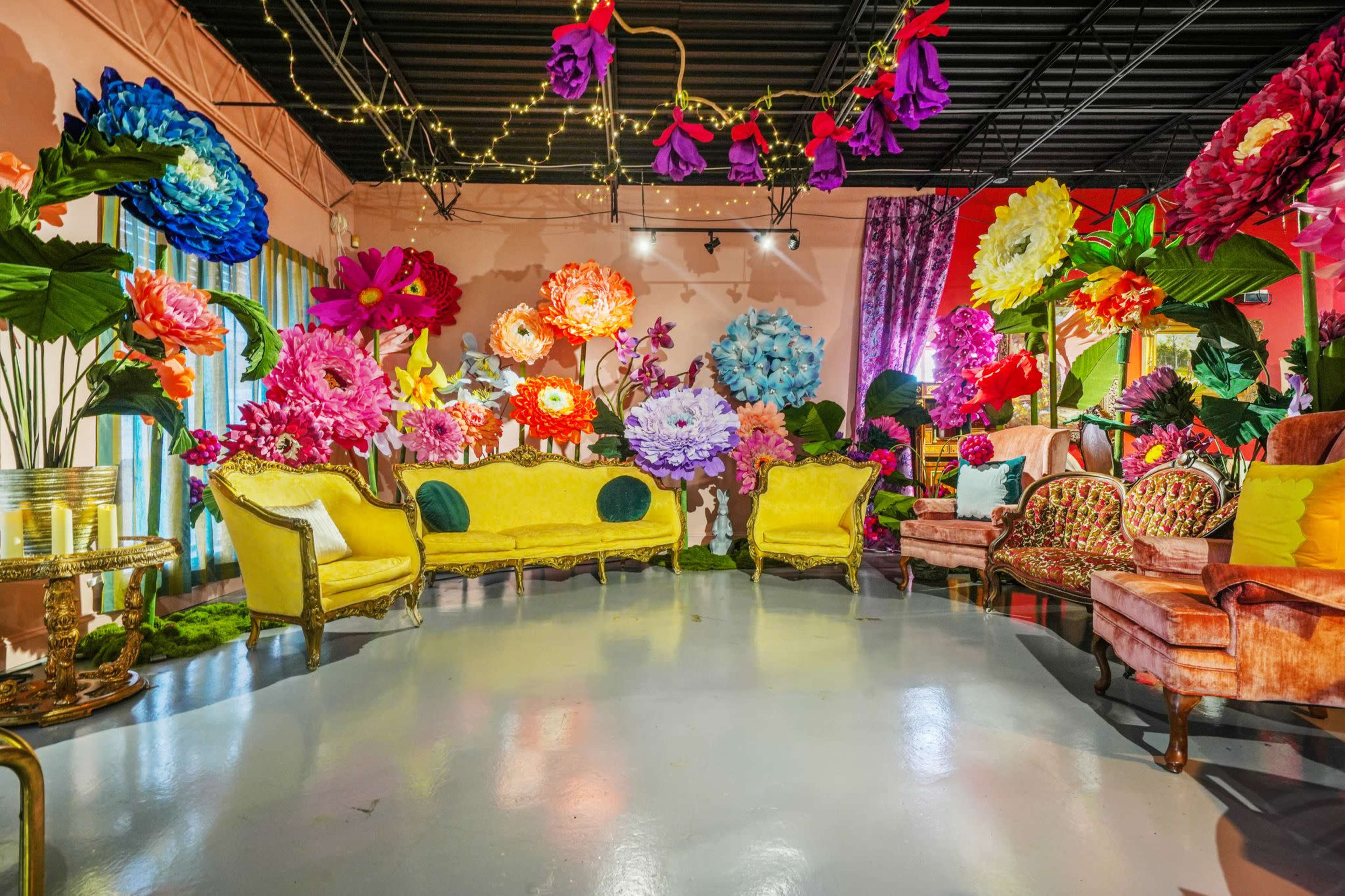 The room features vibrant, oversized paper flowers and colorful furniture arranged around a polished floor, creating a whimsical and artistic atmosphere.