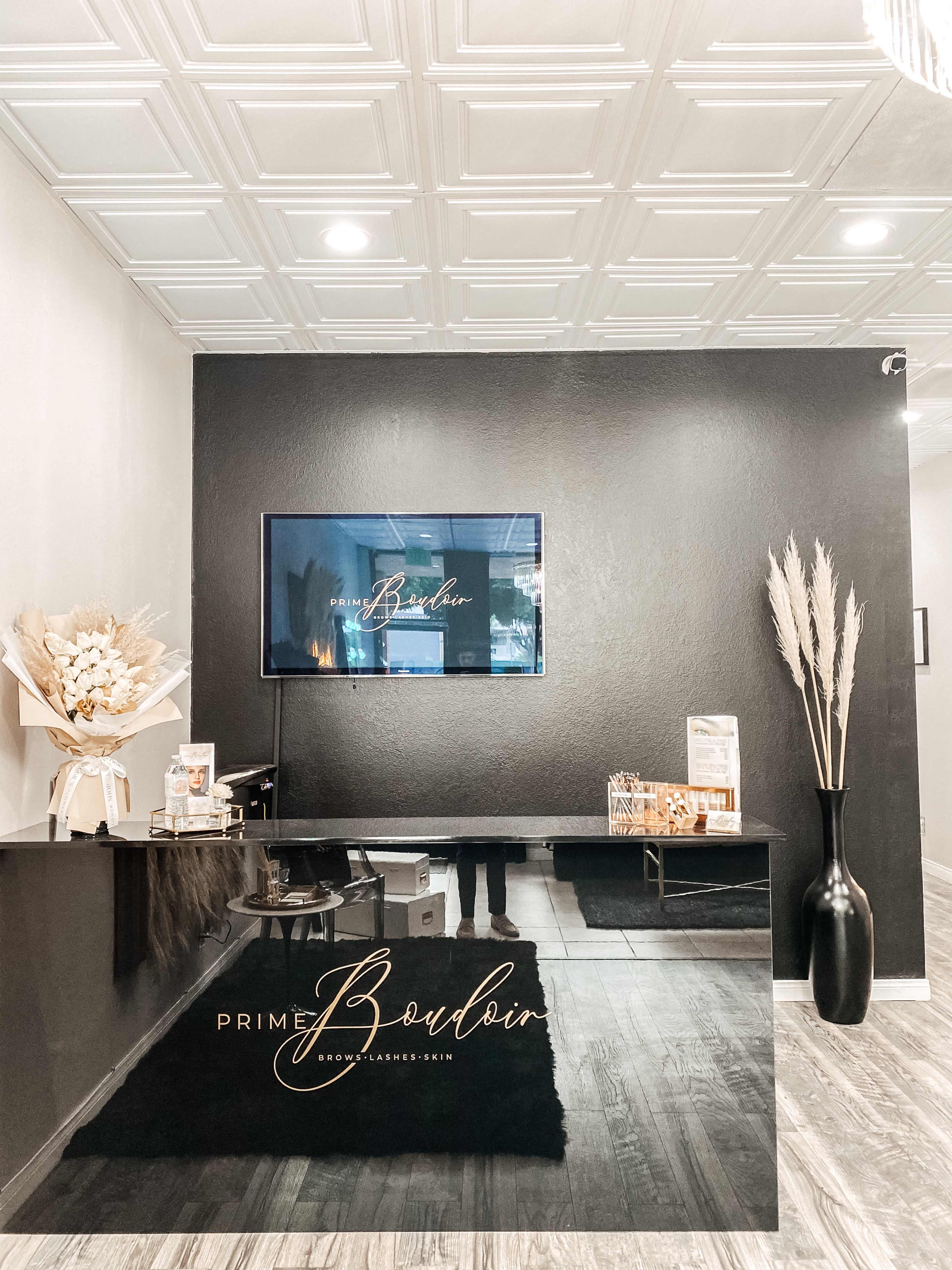 A modern reception area with a dark wall, a sleek black counter, a large TV displaying a logo, and decorative elements including dried plants.
