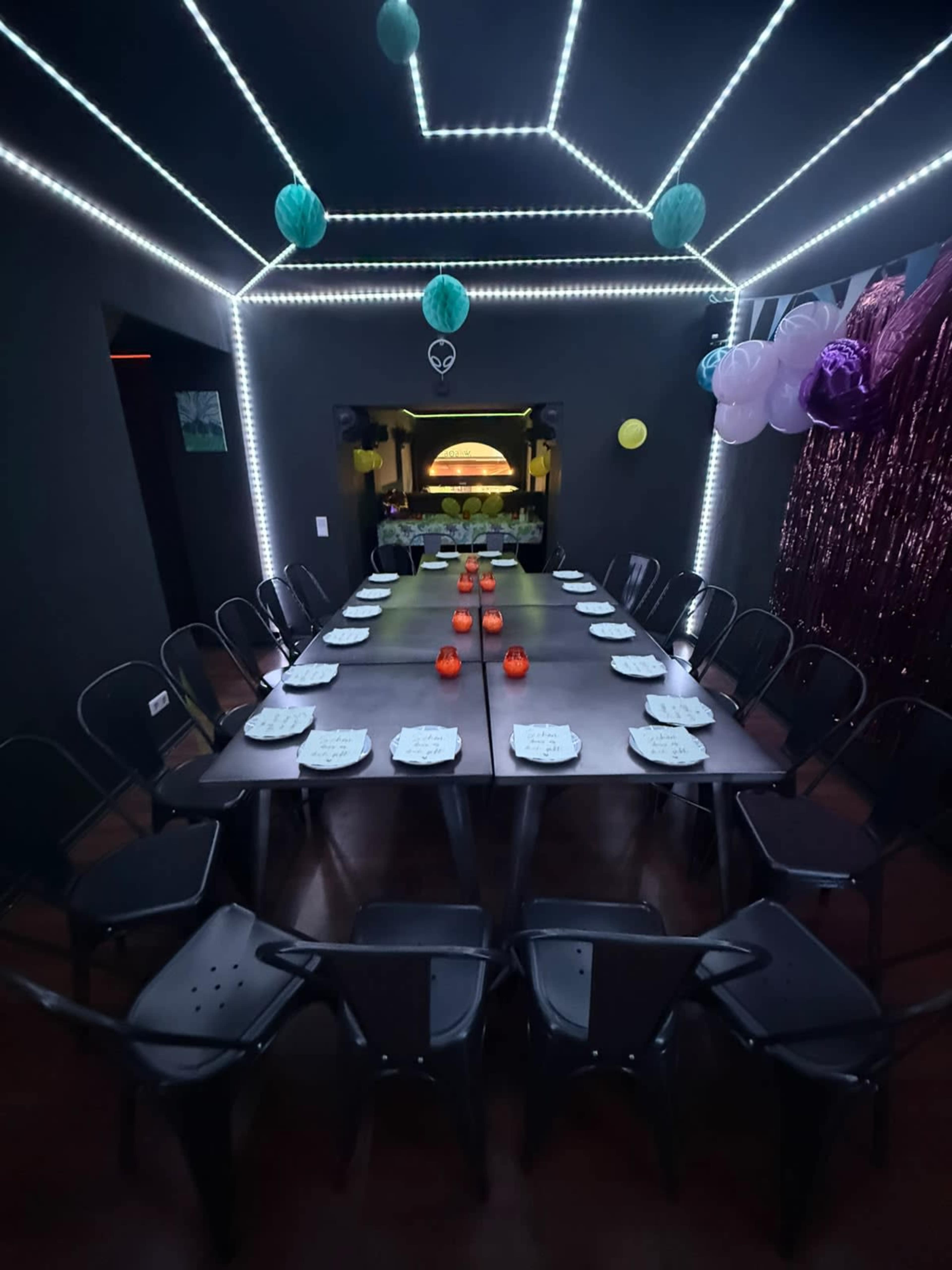 A long dining table is set with plates and decorations in a dimly lit room featuring neon lighting and colorful party decorations.