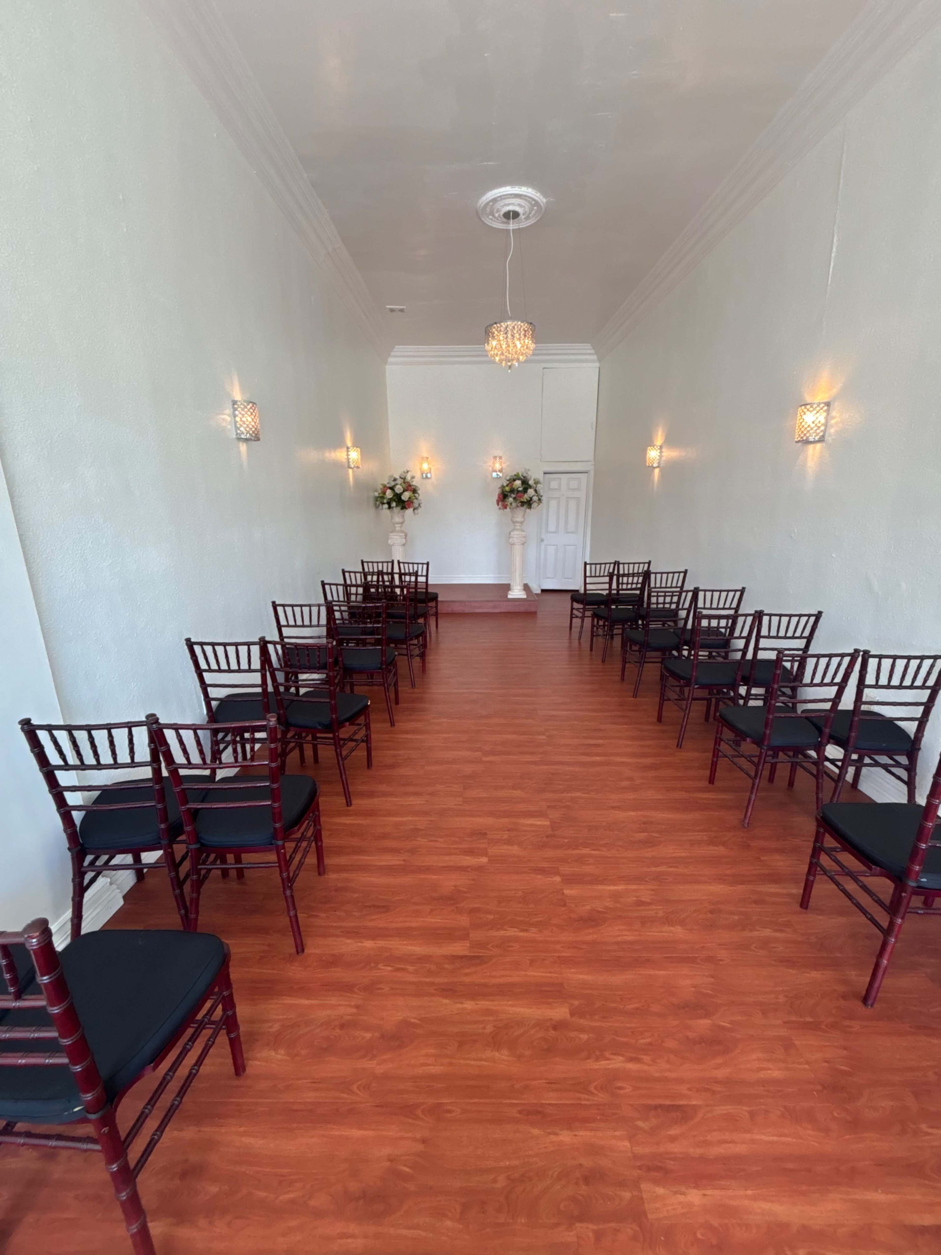 The image shows a narrow indoor venue with wooden flooring, lined with chairs on either side, and floral arrangements at the front.