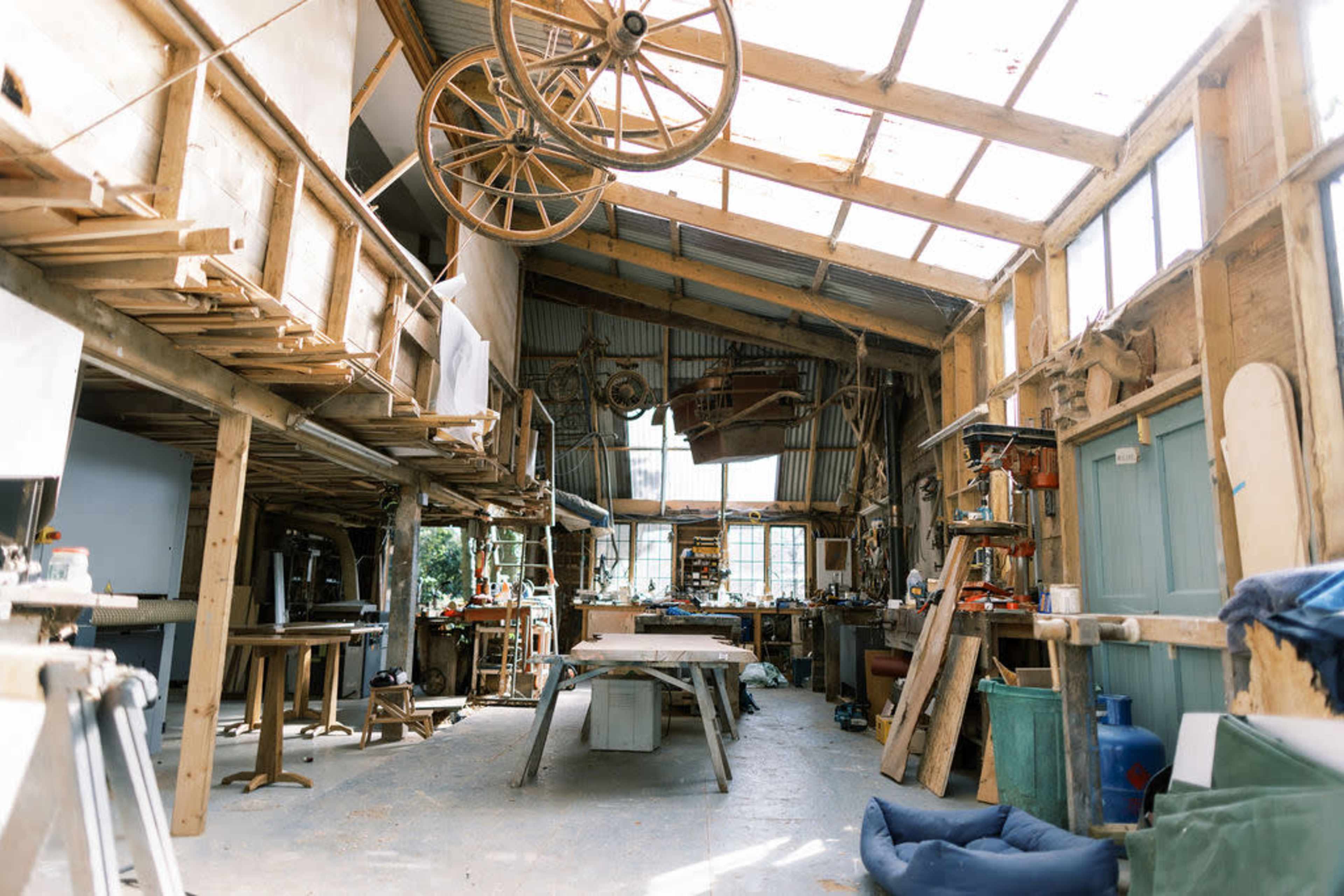A spacious workshop with wooden beams, tools, and equipment scattered throughout, illuminated by natural light coming from large windows.