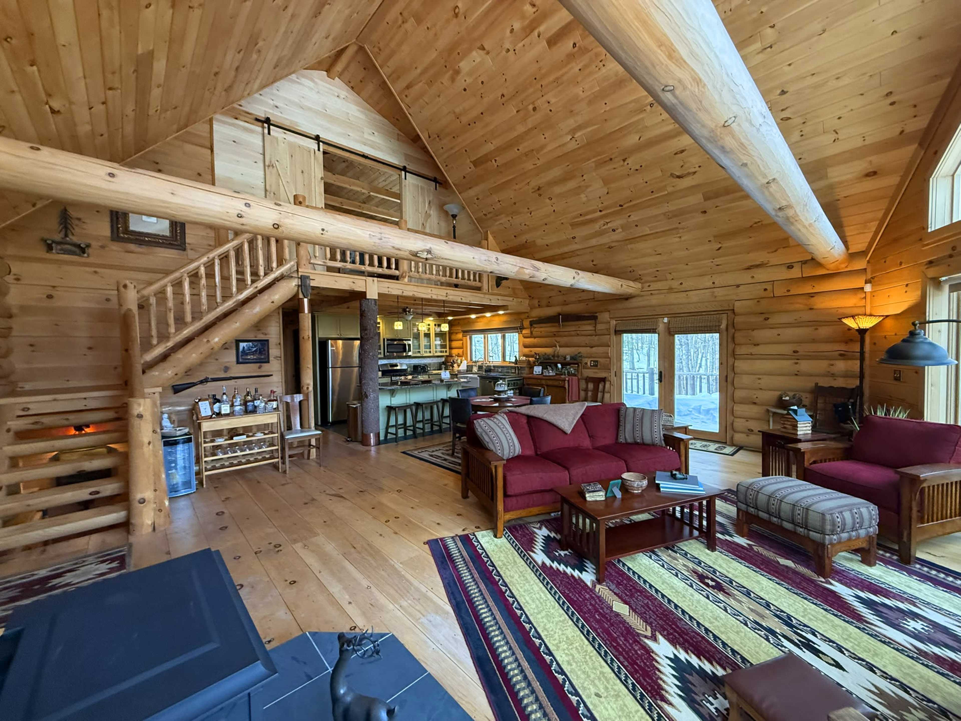 The image shows a spacious wooden cabin interior with a high ceiling, featuring a living area with red sofas, a staircase, and a kitchen in the background.