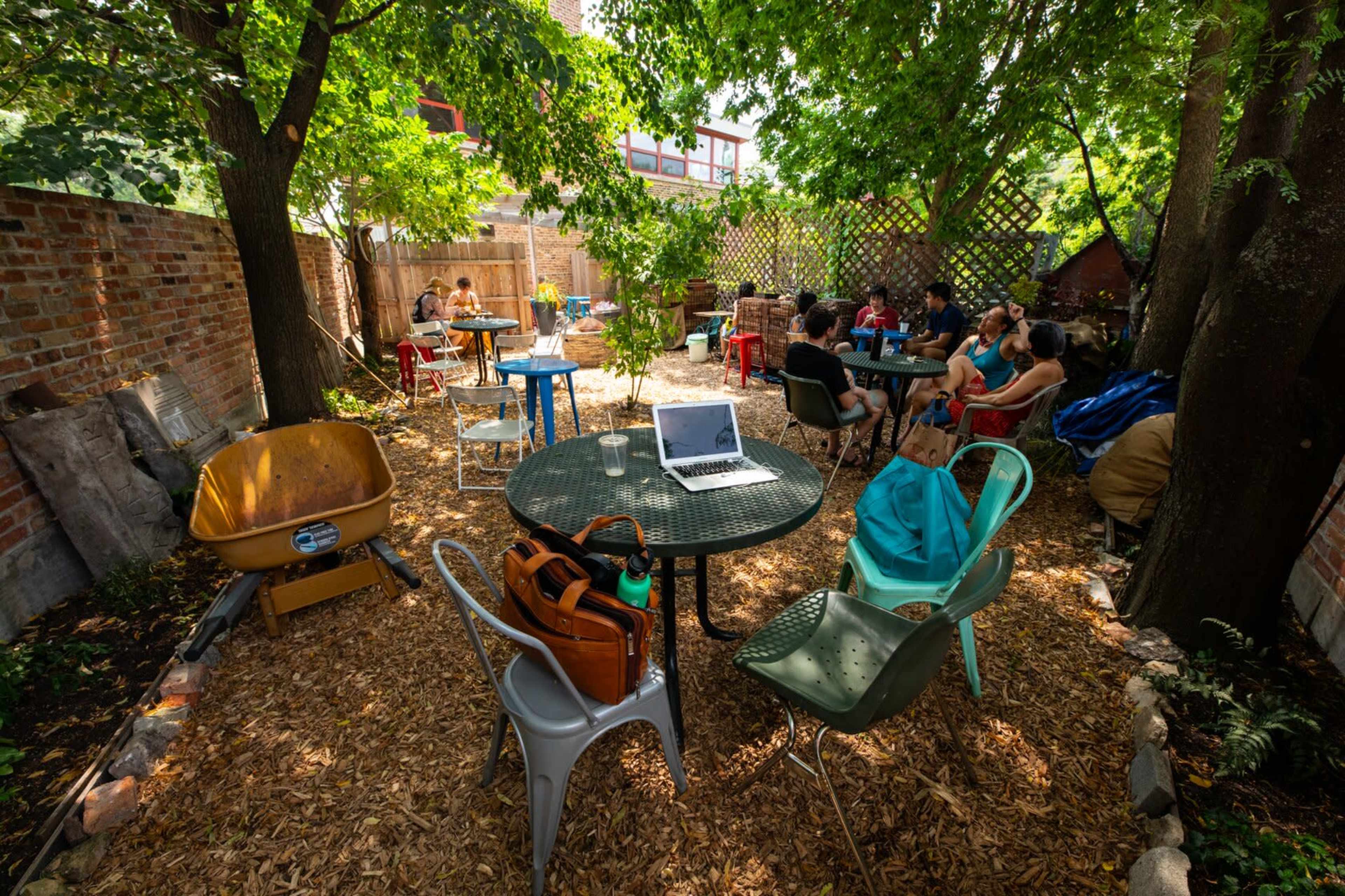 A shaded backyard space features people sitting at various tables amidst greenery and wood chips, with a laptop on one of the tables.