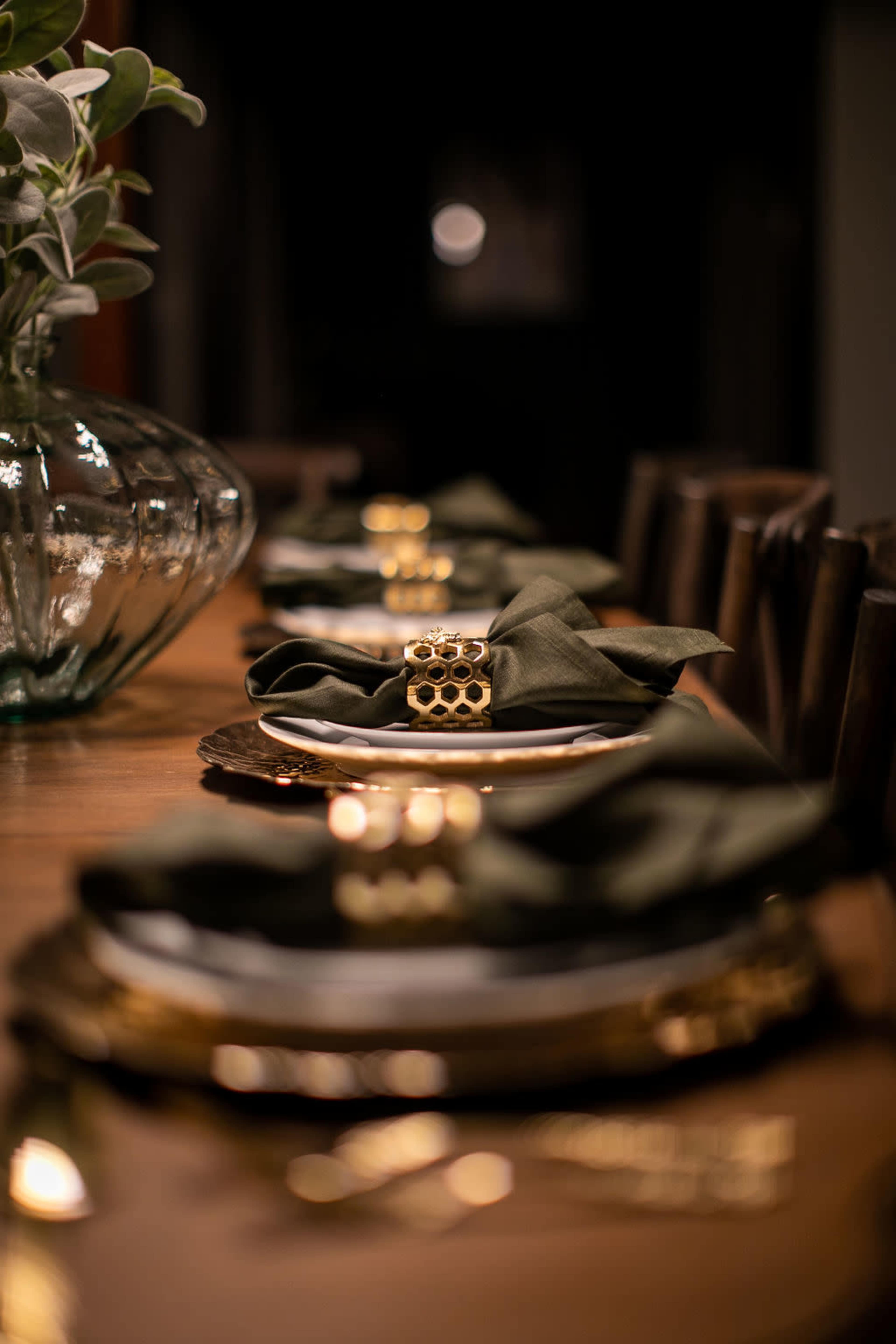 A dining table is set with dark green napkins, decorative napkin rings, and plates arranged on a polished wooden surface, with a glass vase containing greenery in the background.