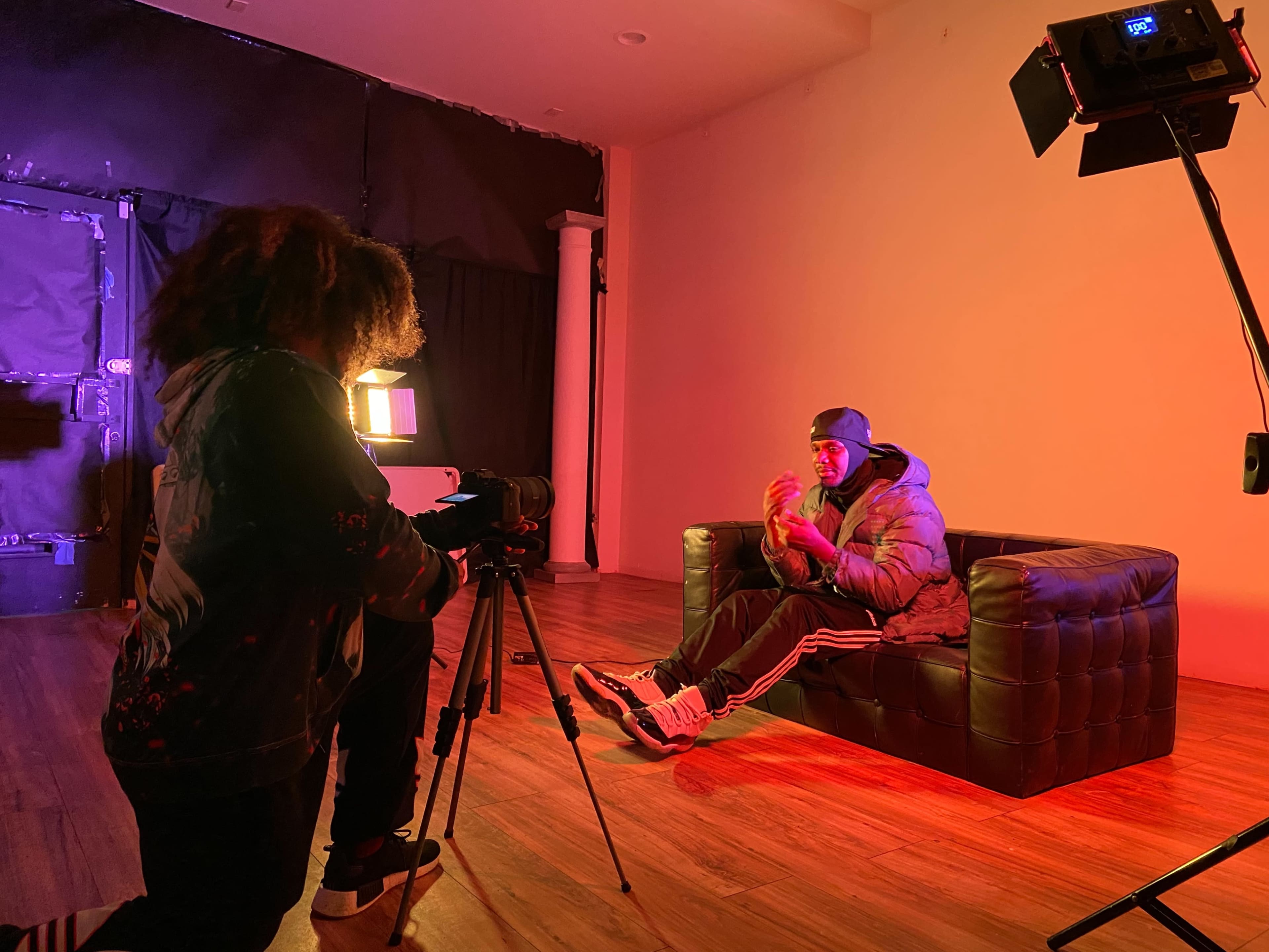 A person sits on a dark leather couch in a dimly lit room while another person operates a camera on a tripod, capturing the scene.