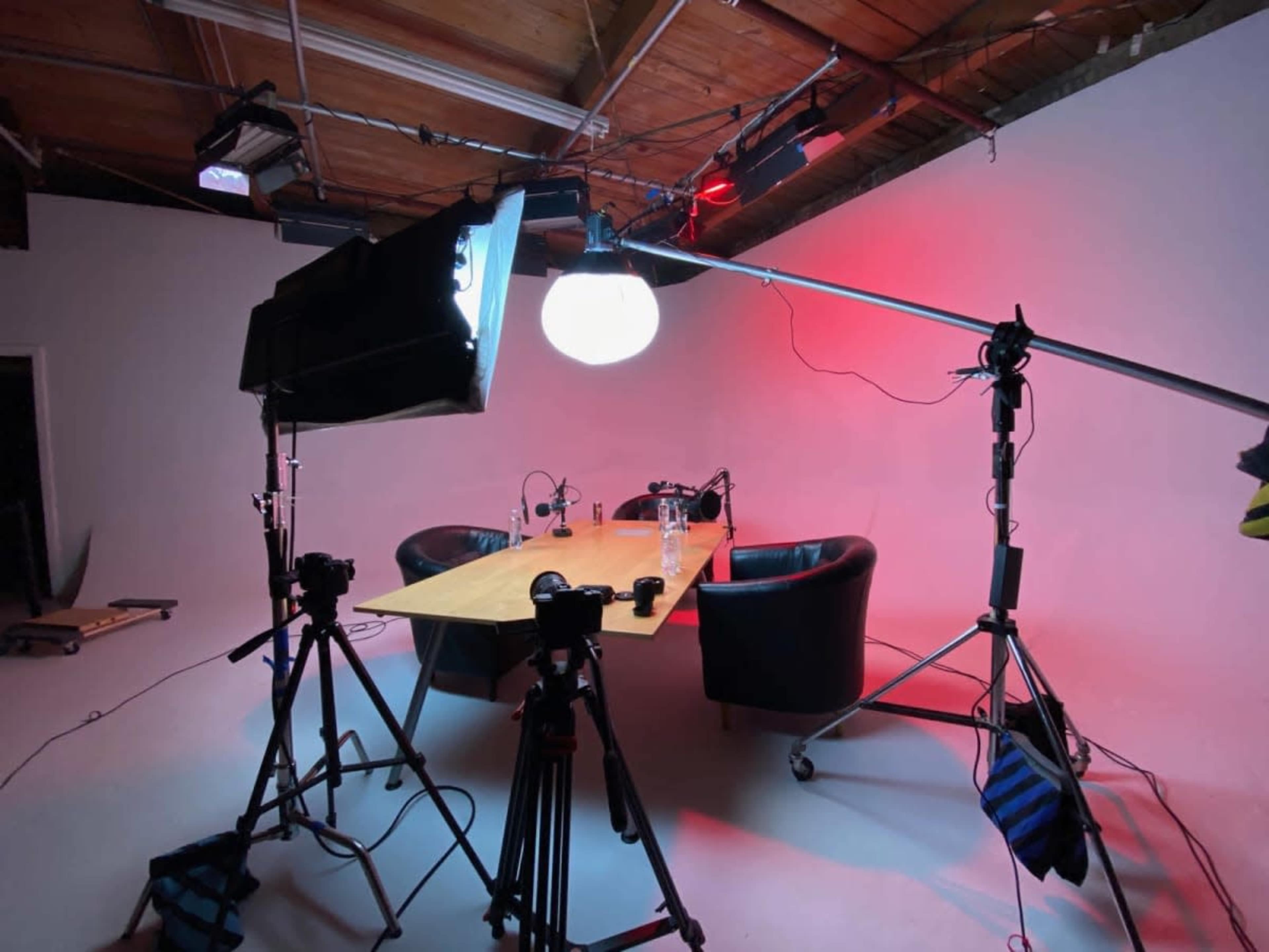 A video recording setup features a table with two chairs, surrounded by lighting equipment and cameras in a studio with a plain background.