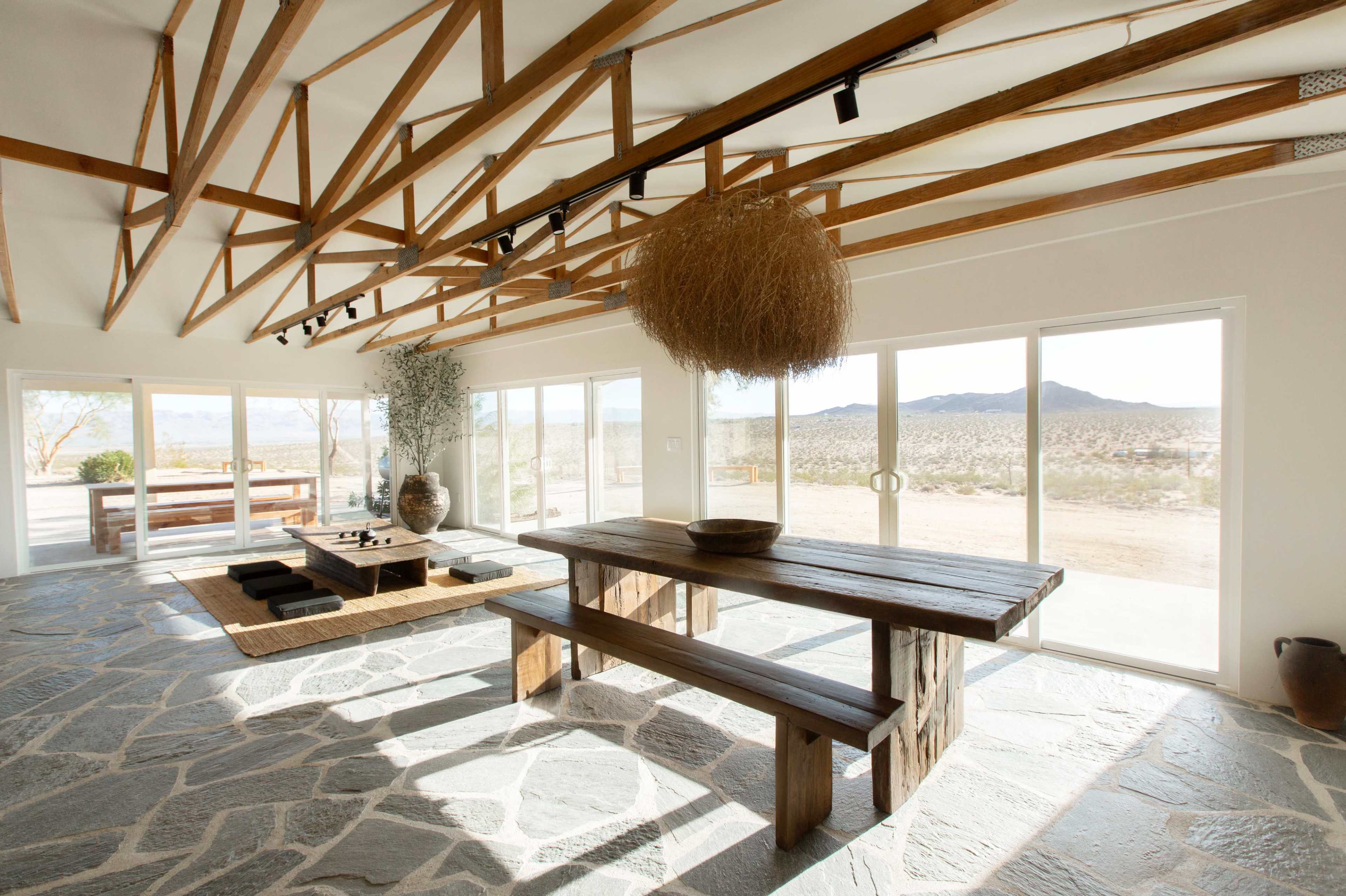 The interior of a spacious room features a wooden table and benches, with large windows showcasing a desert landscape and exposed wooden beams overhead.