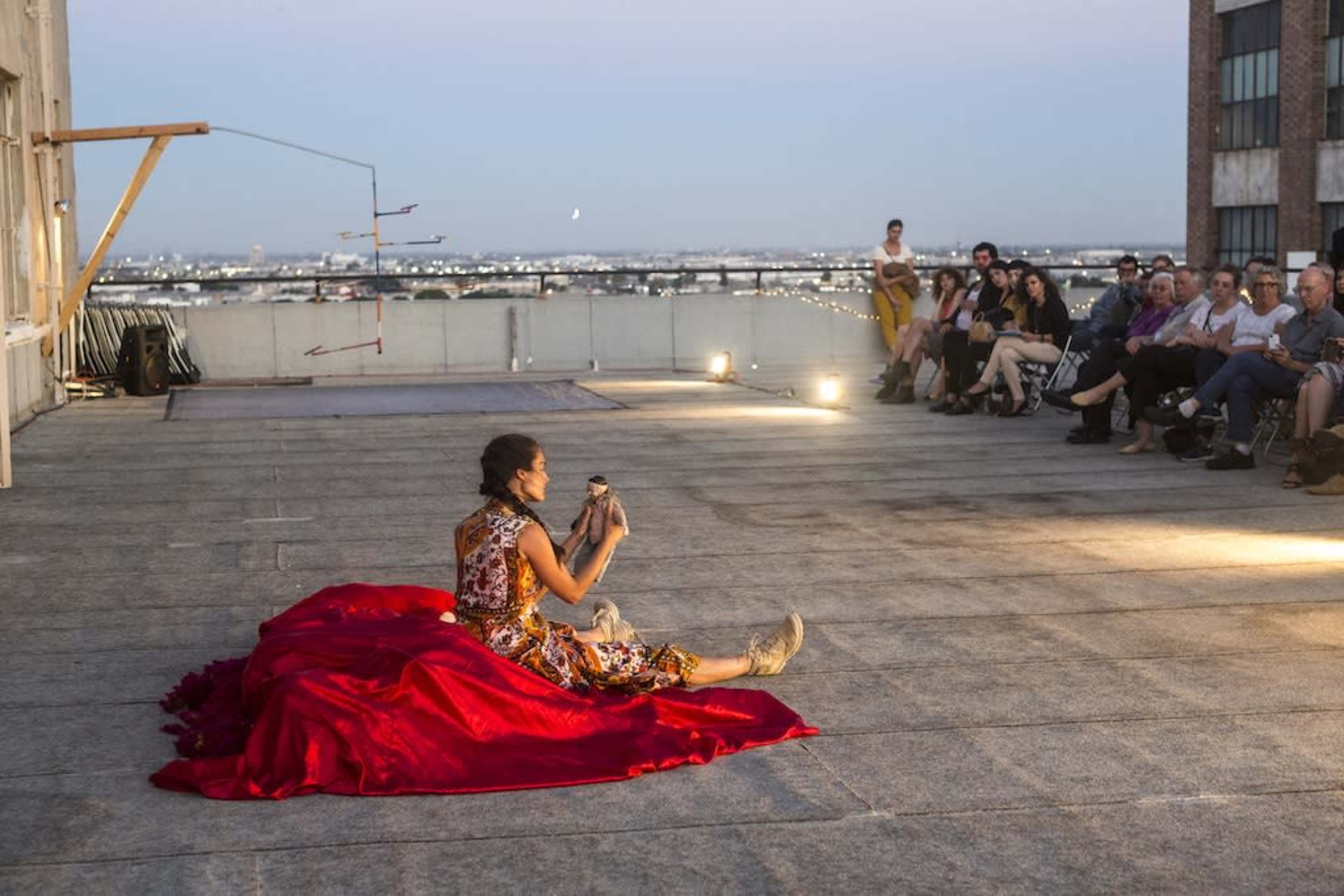 A performer sits on a red fabric while holding a small animal in front of an audience on a rooftop at dusk.