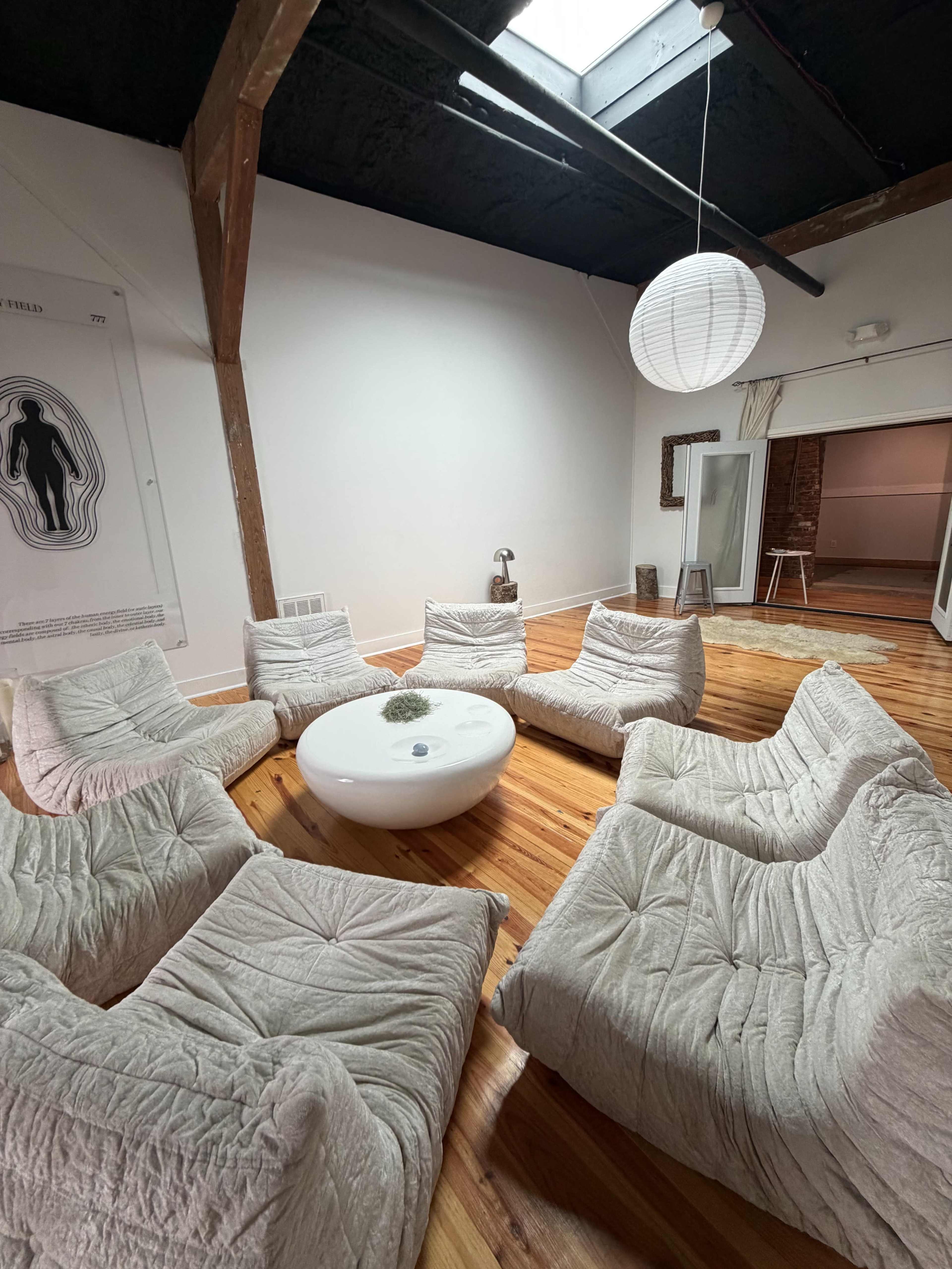 The image shows a spacious, brightly lit room with a circular arrangement of soft, textured seating around a low, round table.