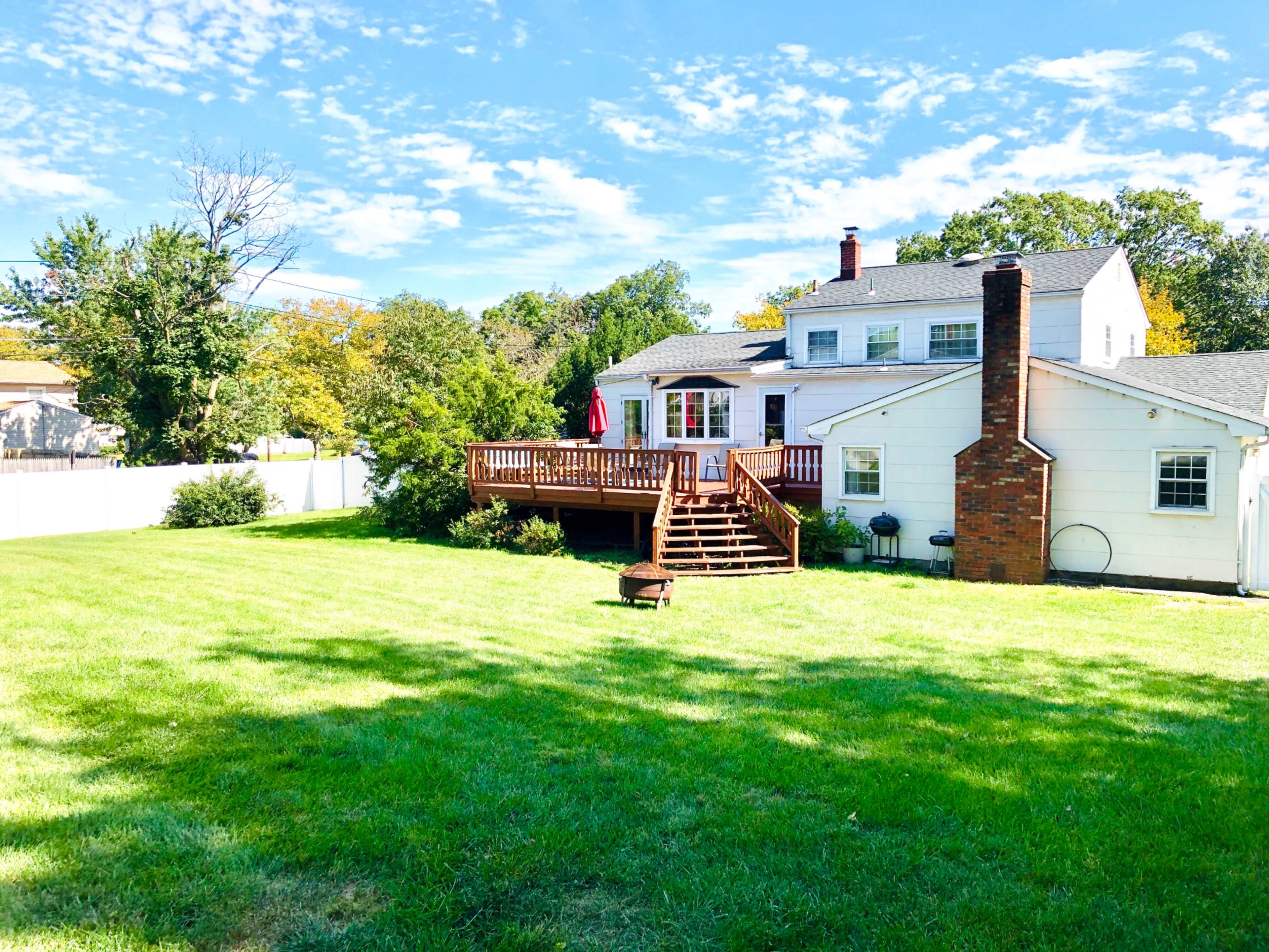 A spacious backyard with a wooden deck attached to a white house, surrounded by green grass and trees under a partly cloudy sky.