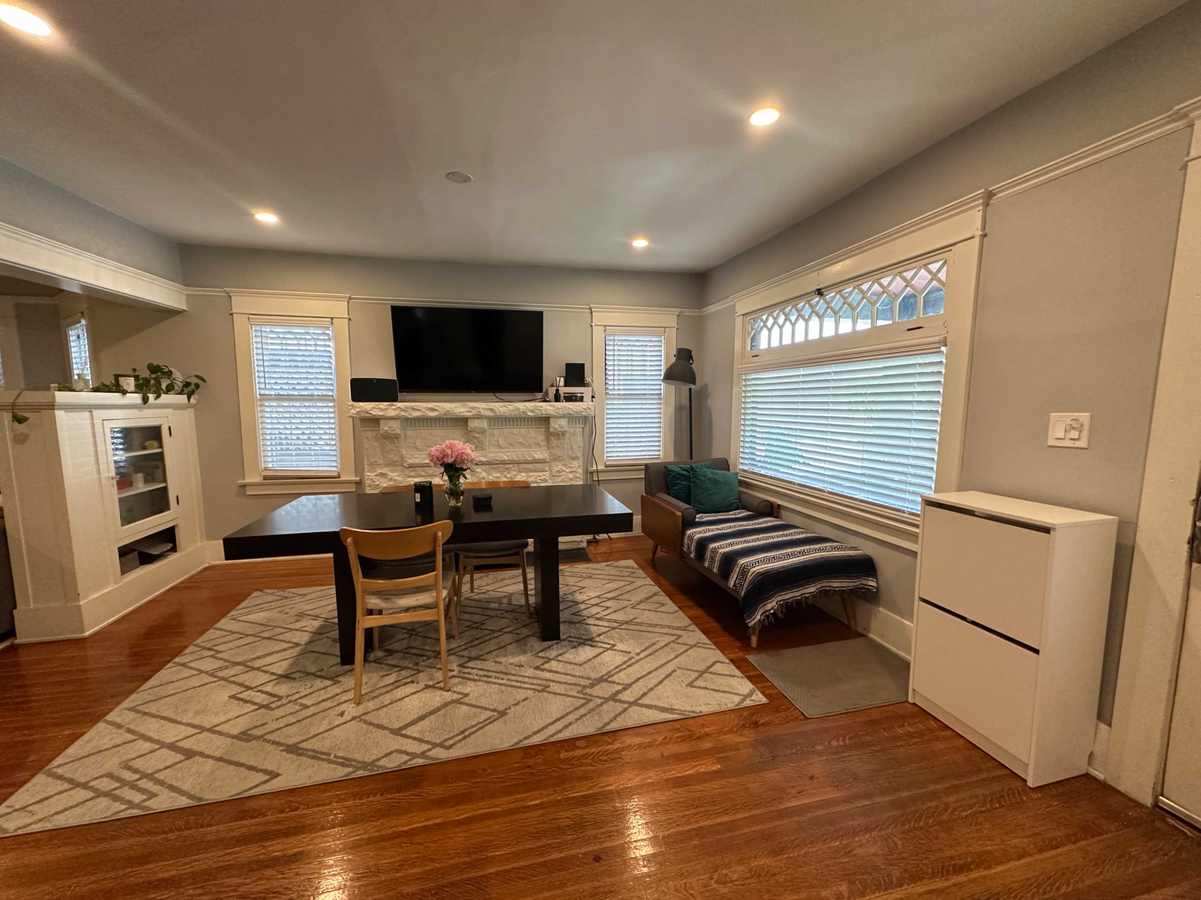 The image shows a modern living room with a dining table, chairs, a white stone fireplace, and large windows with natural light.