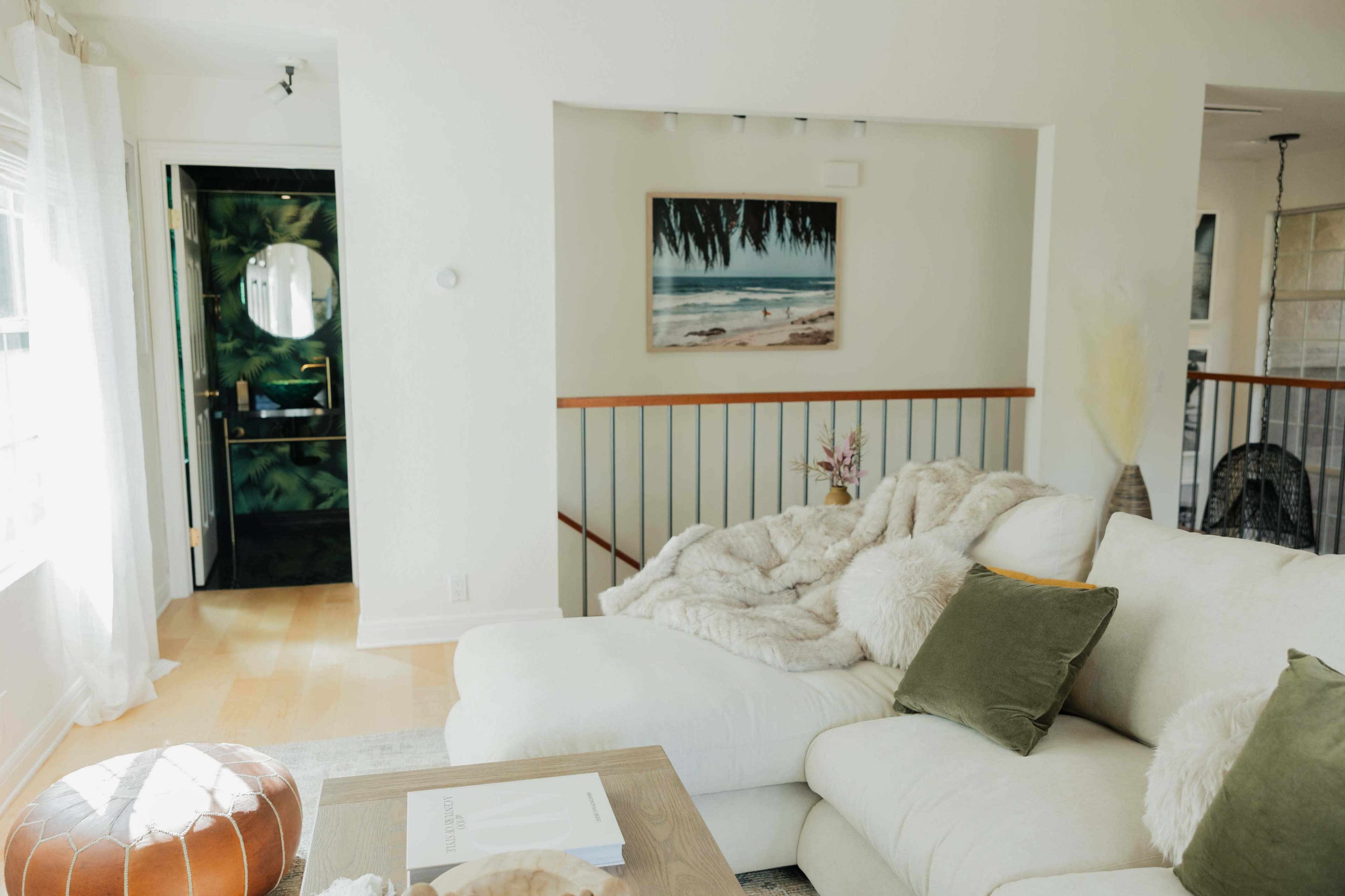 The image shows a living room with a large white sofa, a textured throw, and a wooden coffee table, next to a balcony overlooking a green door and coastal artwork on the wall.