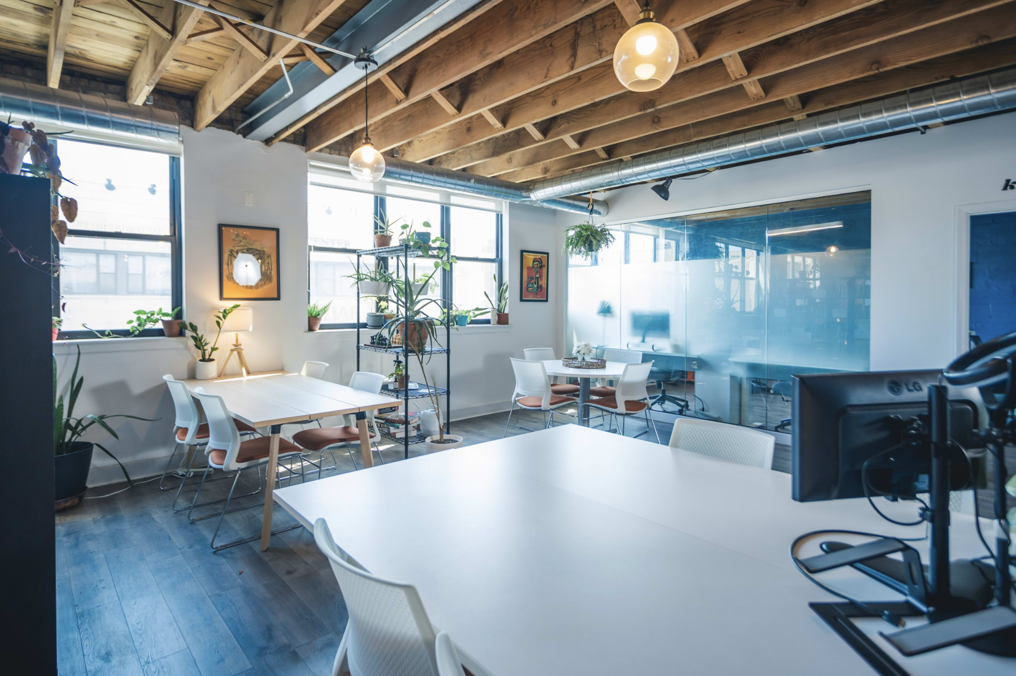 A modern office space with wooden beams, multiple workstations, plants, and large windows allowing natural light.