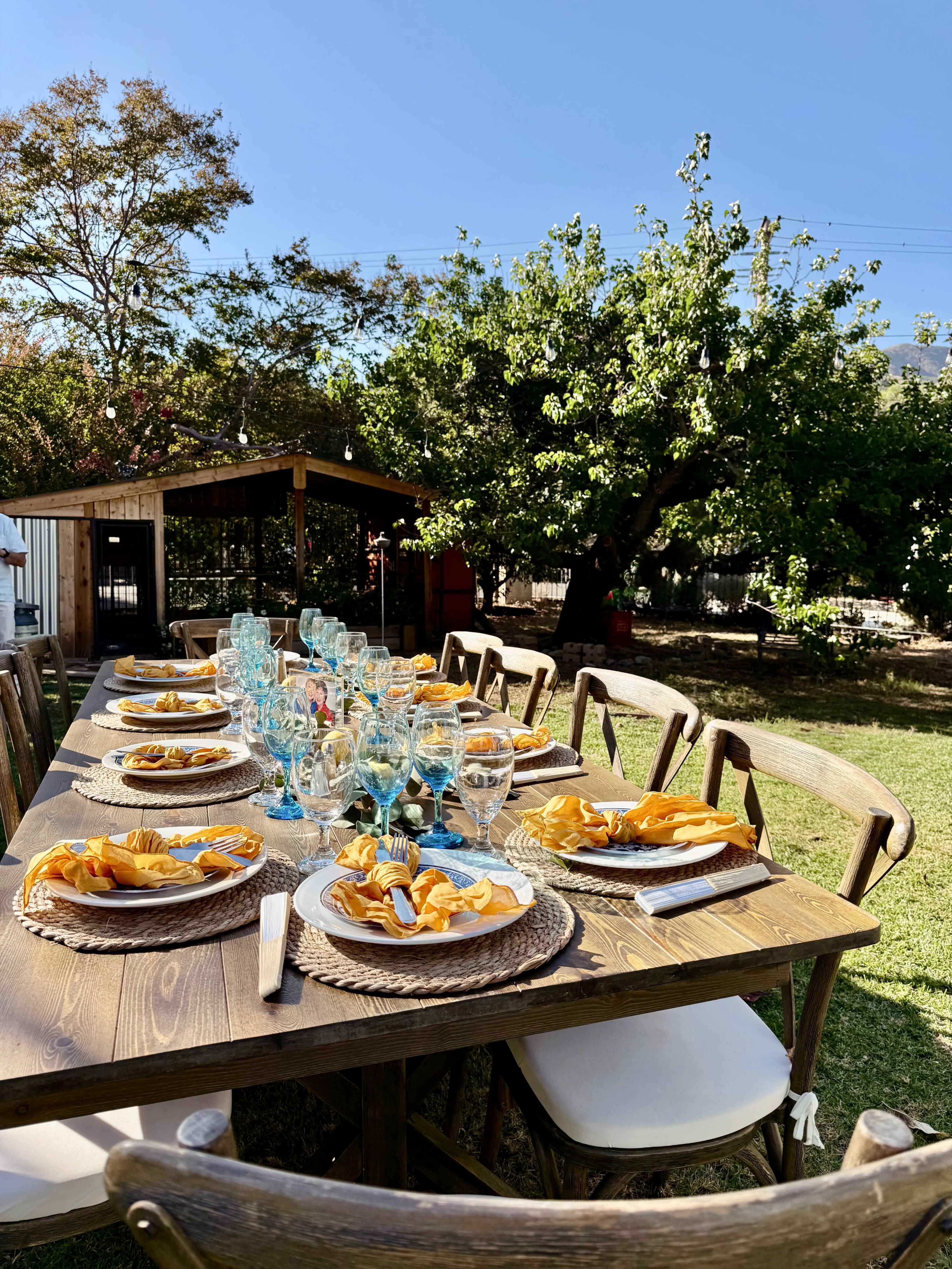A long wooden table is set outdoors with neatly arranged plates of food and glasses, surrounded by greenery and a small building in the background.