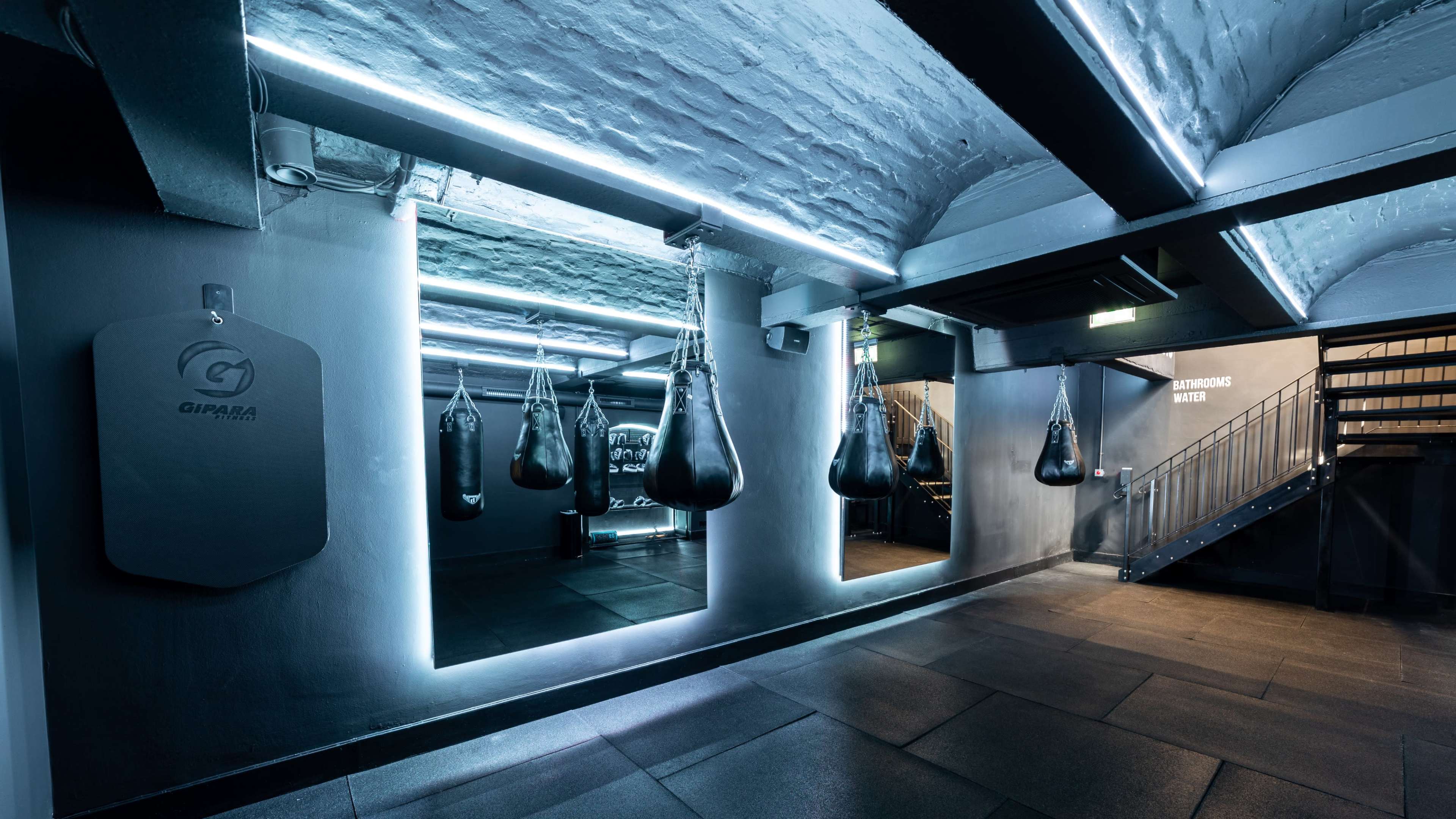 The image shows a modern gym interior featuring several hanging punching bags, illuminated mirrors, and a staircase in a dimly lit environment.