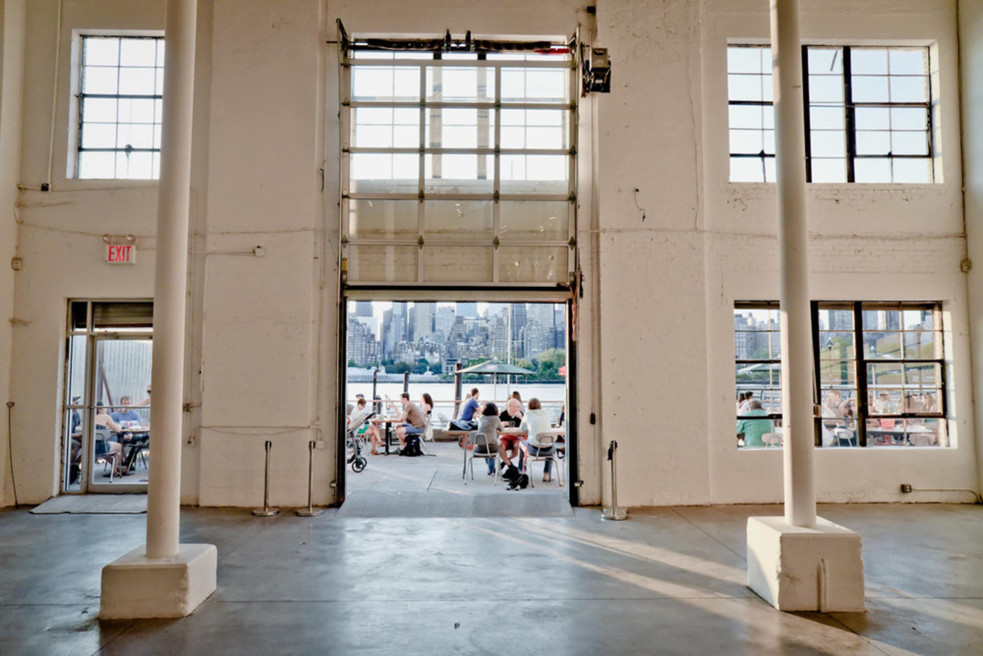 An open industrial-style entrance with large glass doors, revealing a view of a bustling outdoor area filled with people and the city skyline in the background.