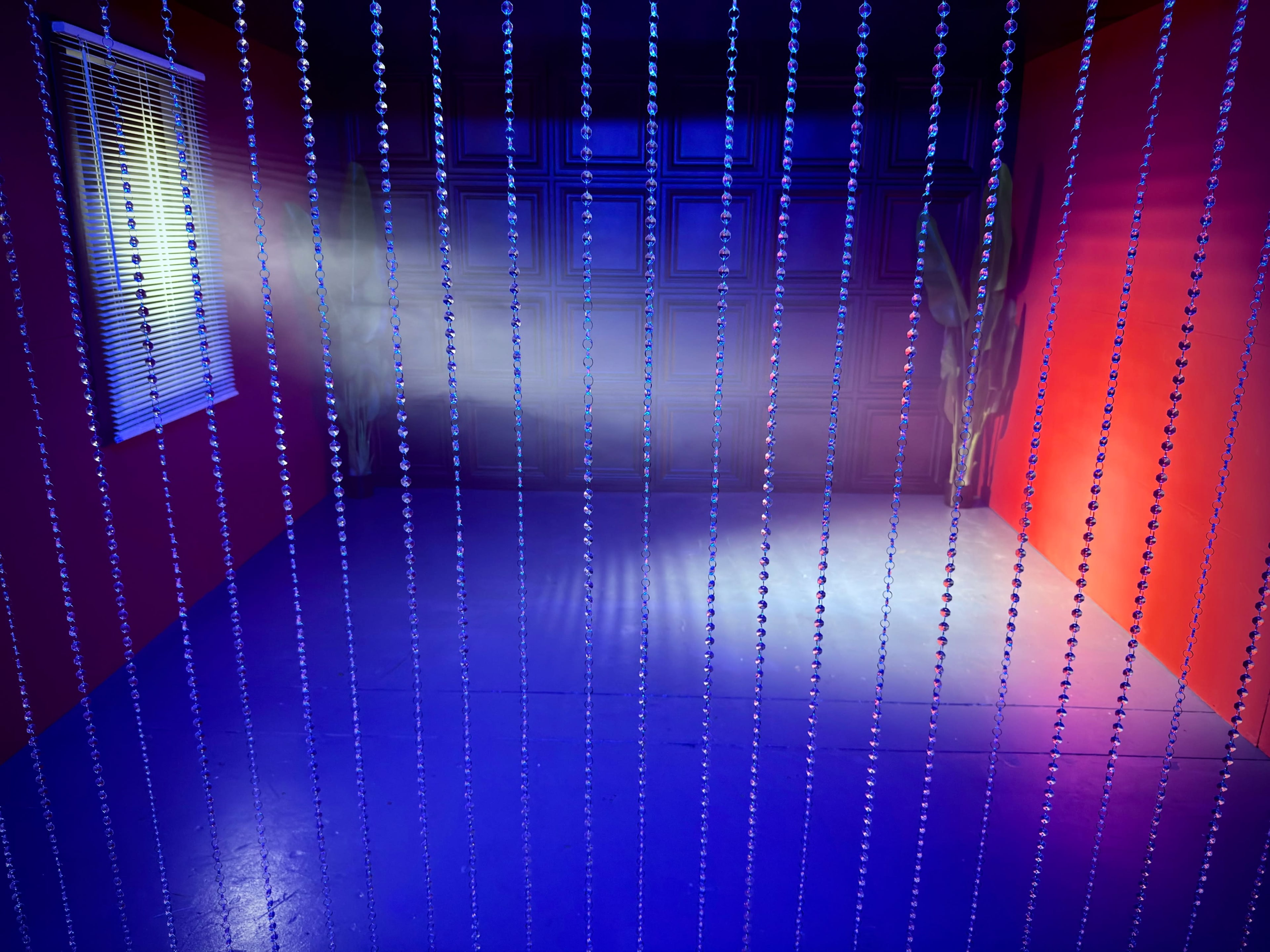 A brightly lit room features a wall with red paint, a window with blinds, and a beaded curtain separating the space.