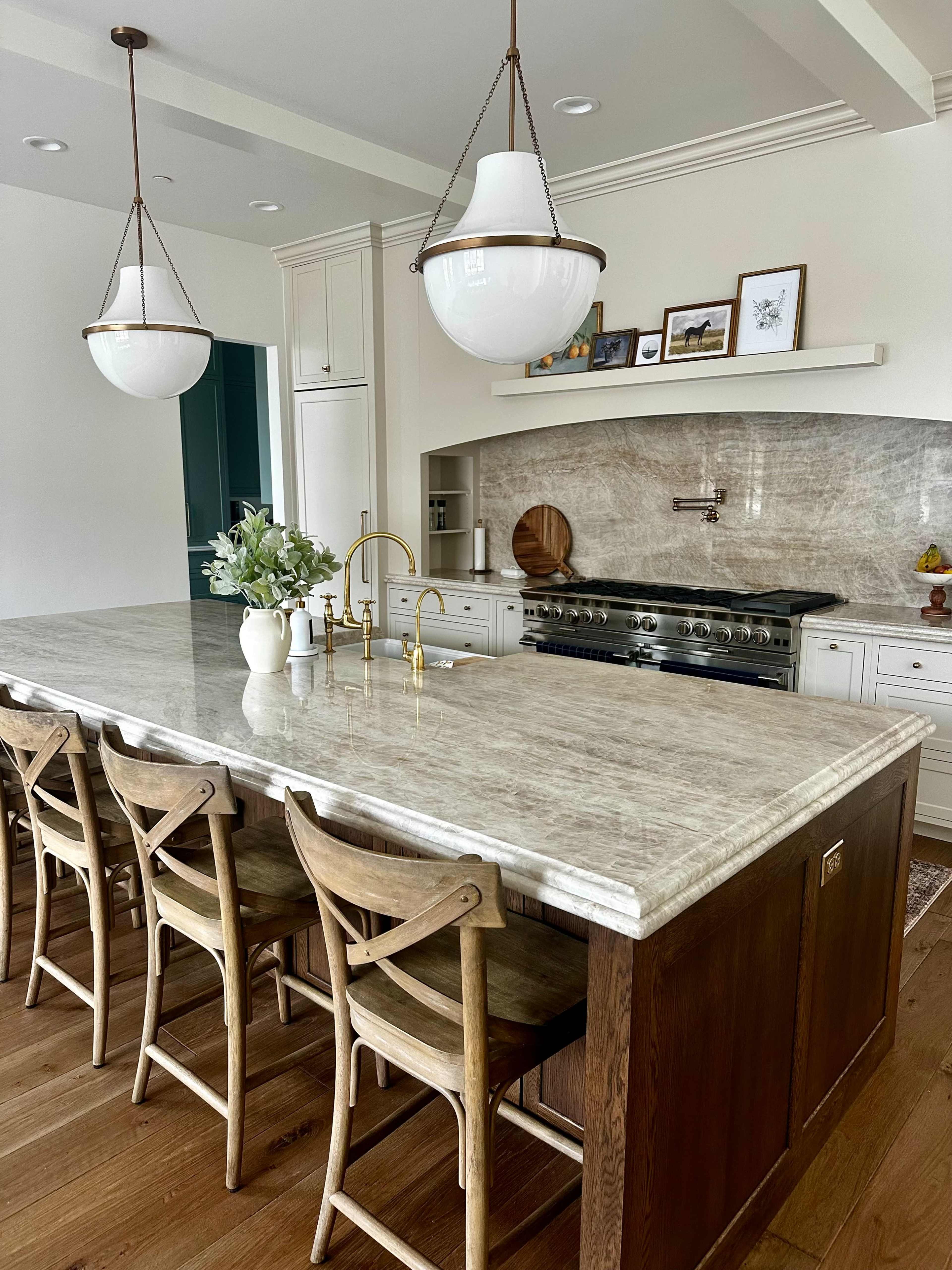 The image features a modern kitchen with a large marble island, wooden stools, and pendant lighting above a countertop, along with a gas stove and decorative items on the shelves.