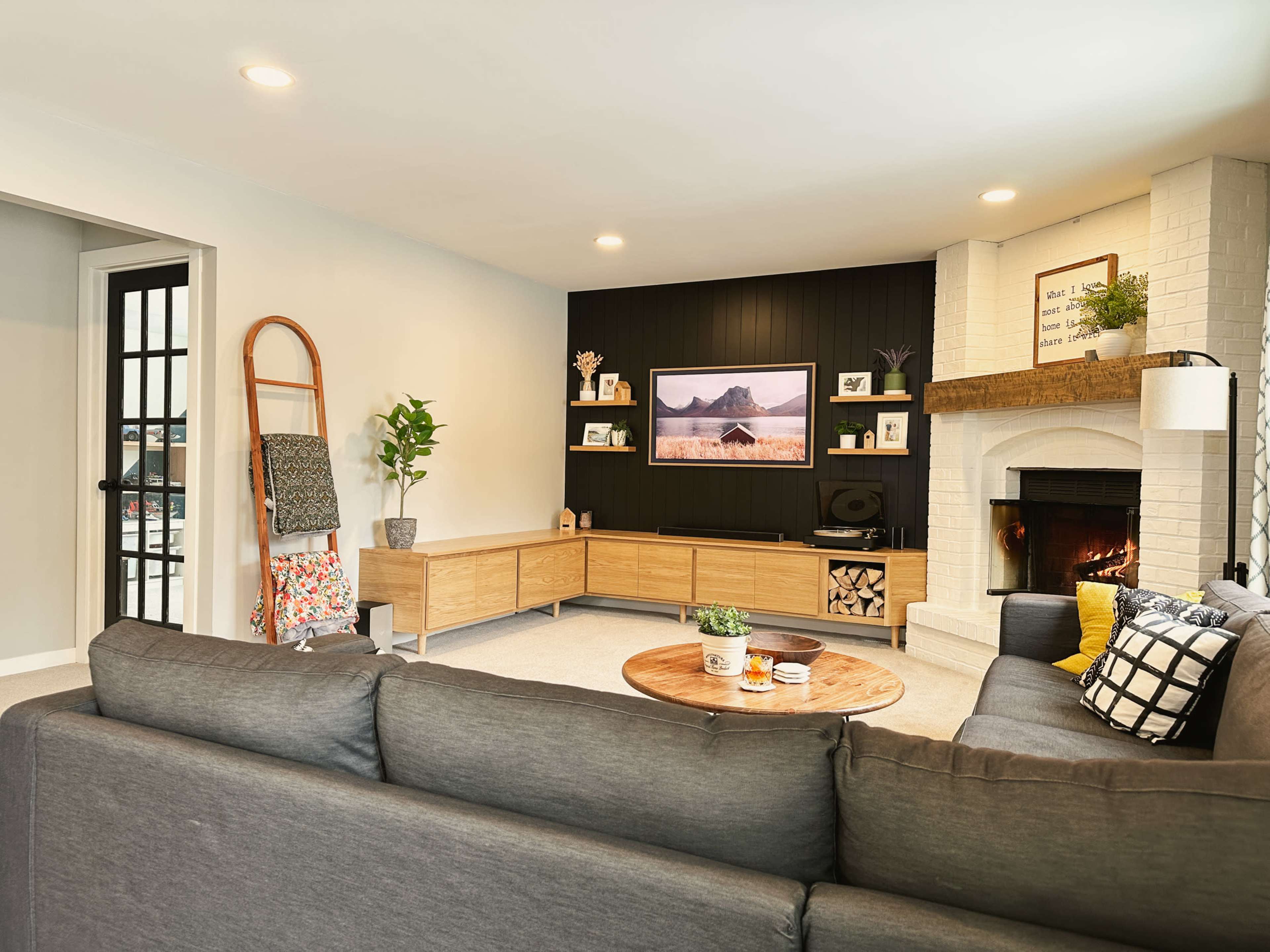 The image shows a modern living room with a gray sectional sofa, a circular coffee table, a TV mounted on the wall, and a fireplace surrounded by light-colored brick.
