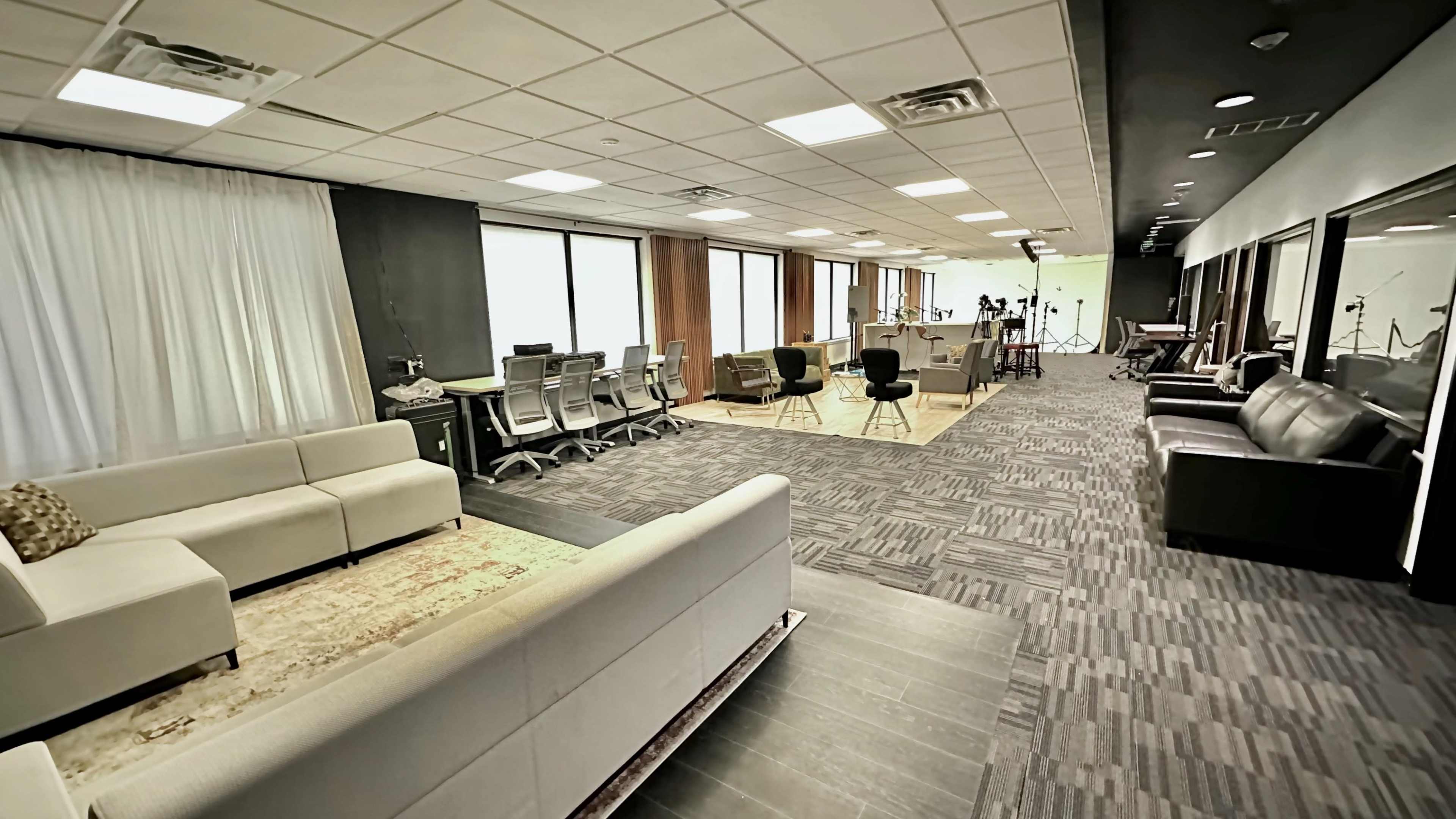 The image depicts a spacious office area featuring seating arrangements, desks, and large windows allowing natural light to enter.