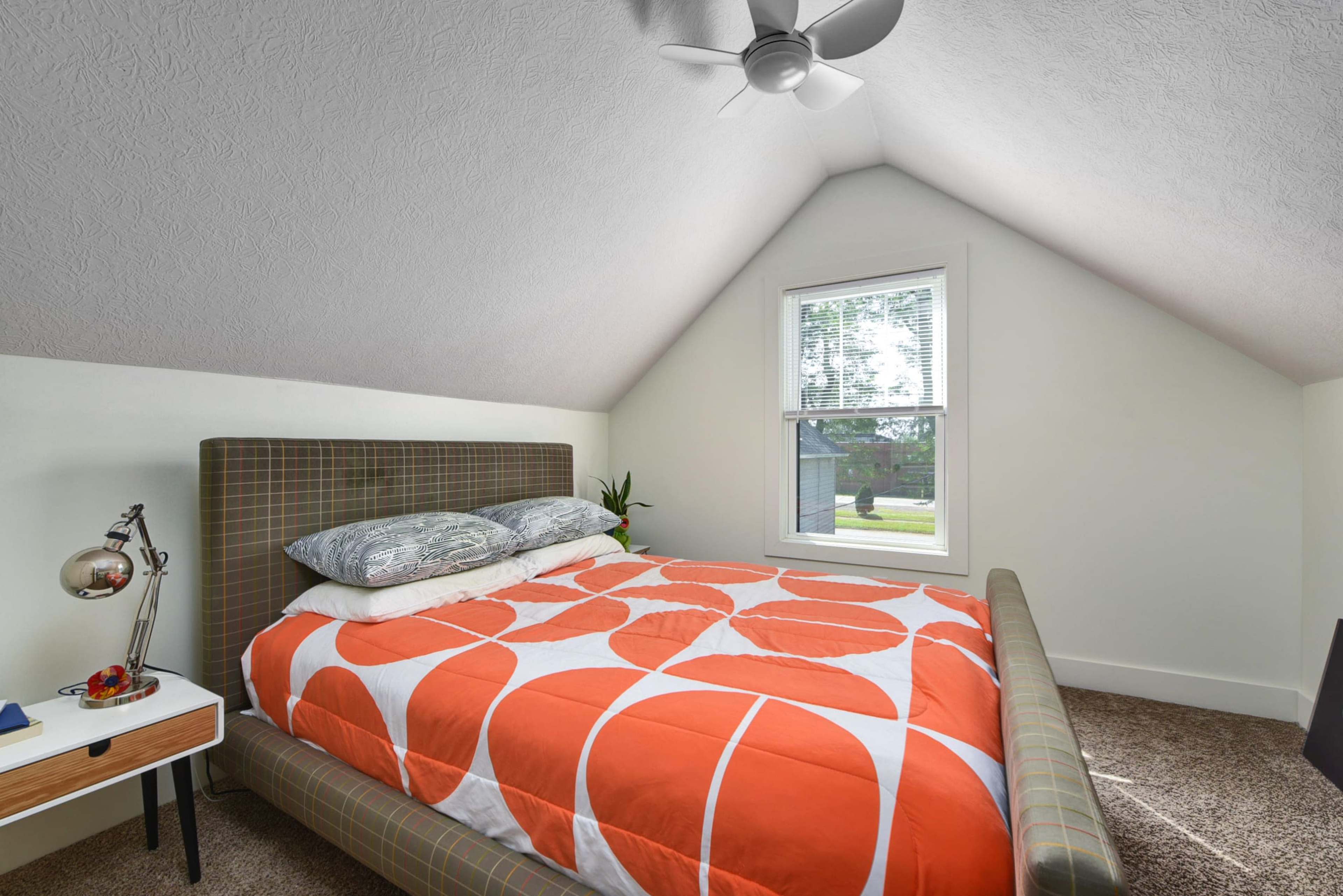 A cozy bedroom with a sloped ceiling, featuring a bed with an orange and white patterned comforter, a bedside table with a lamp, and a window that provides natural light.