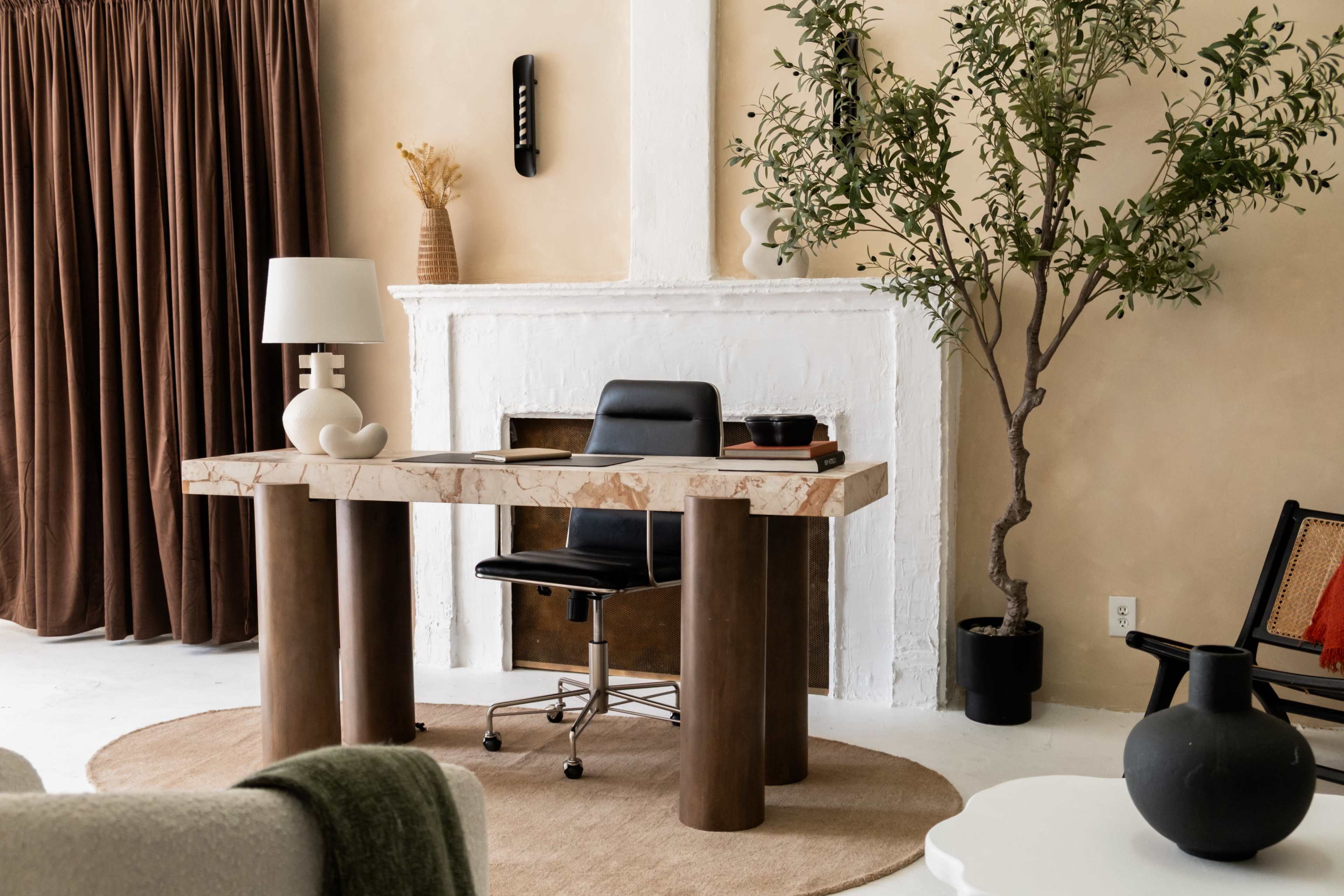 A modern workspace features a unique marble-top desk, a black office chair, and a lamp, set against a backdrop of a textured wall and decorative elements like a potted plant and a fireplace.