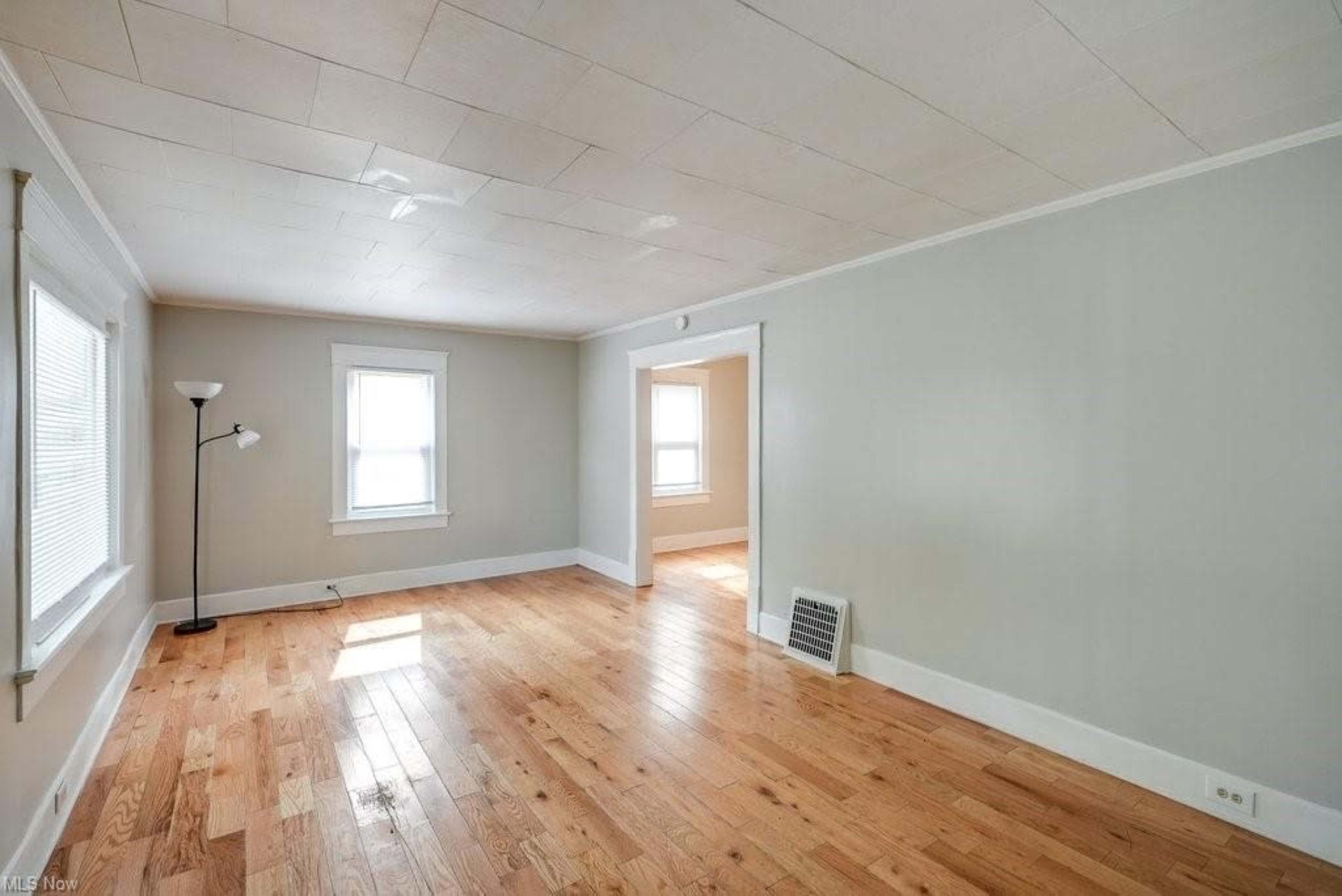 A vacant room with light-colored walls and hardwood flooring, featuring windows on two sides and a standing lamp in the corner.