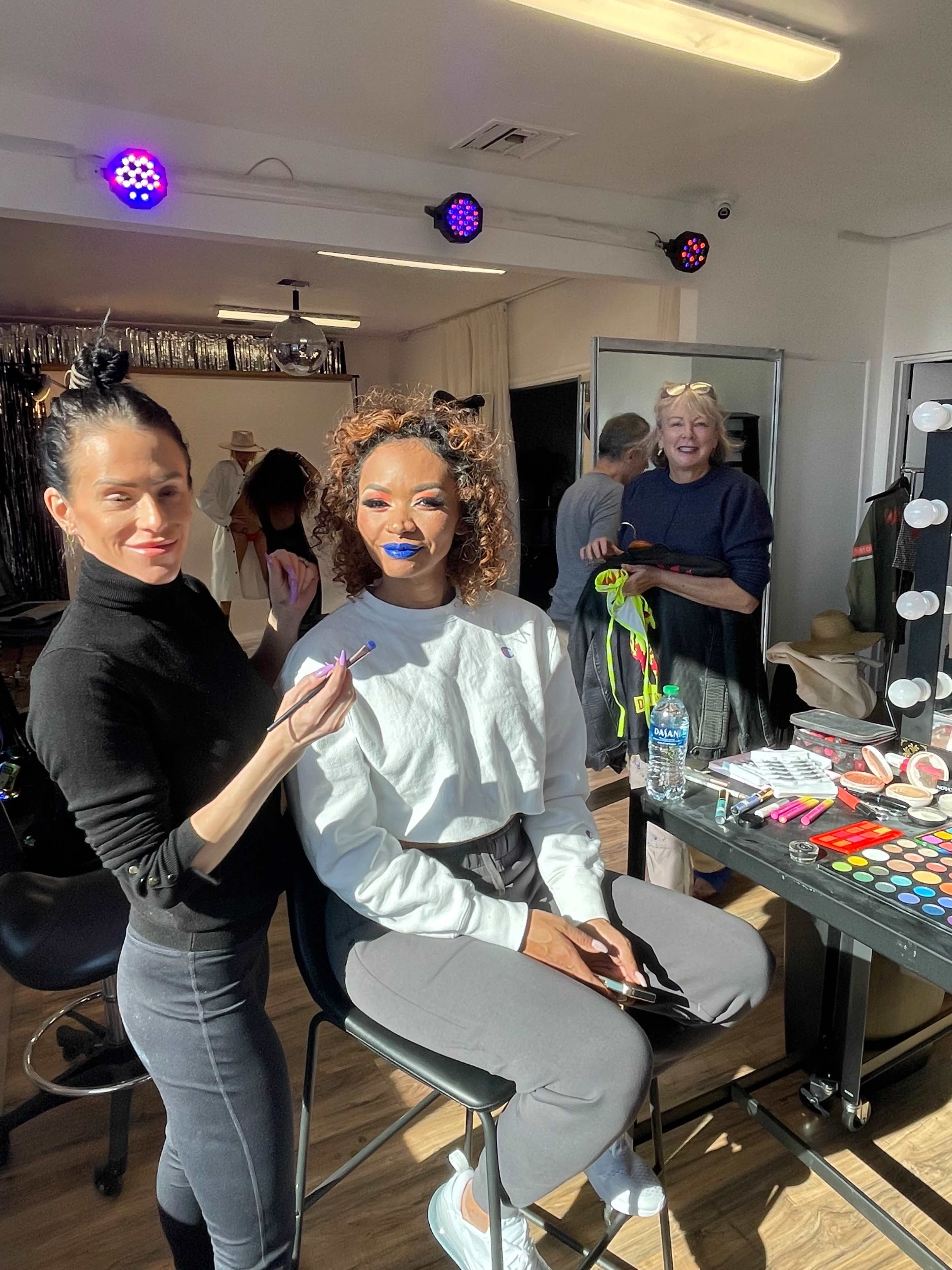 A makeup artist applies lipstick to a woman seated in a stylistic studio, with another person smiling in the background.