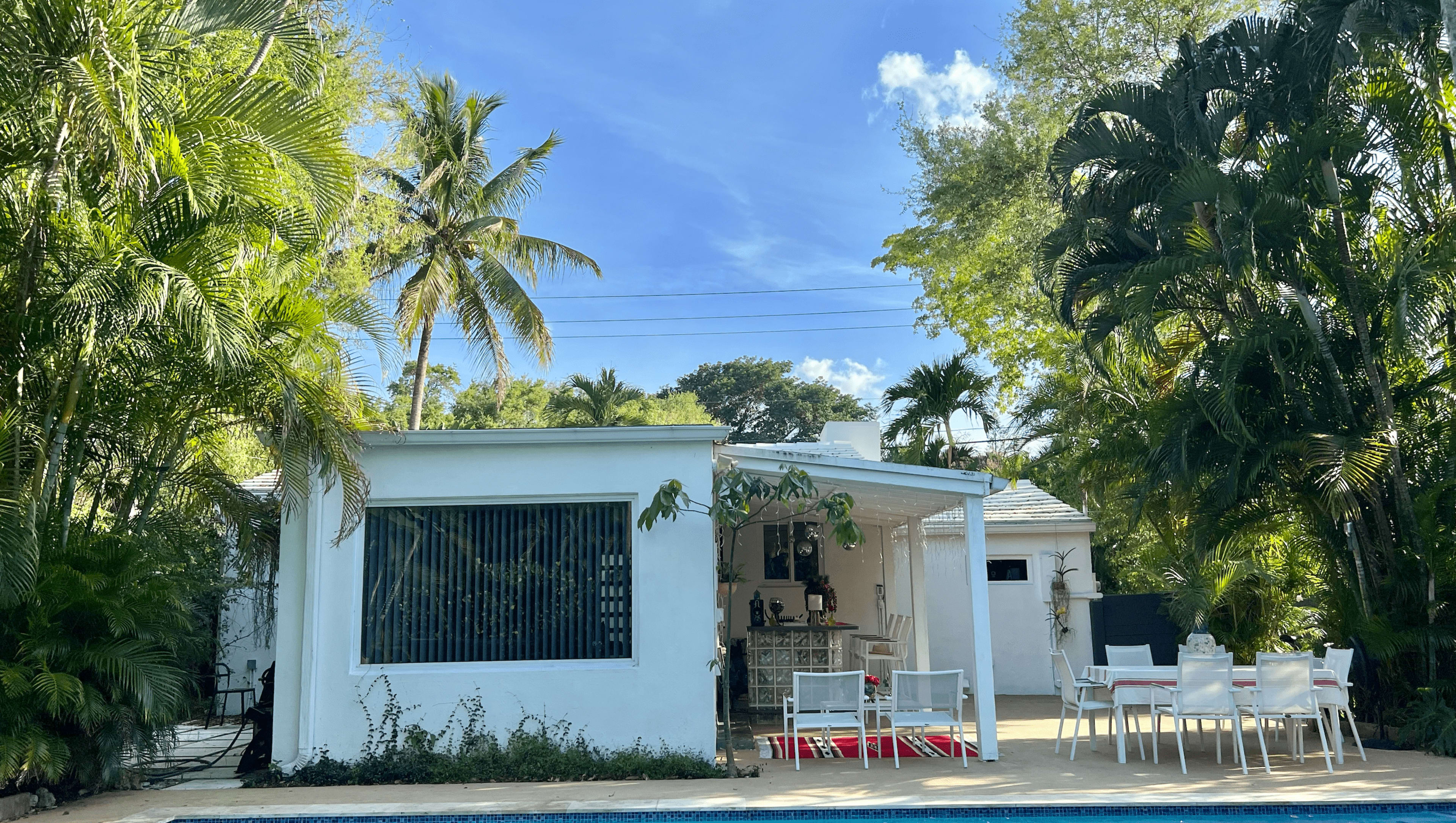 A modern white house with a patio and pool is surrounded by tall palm trees and lush greenery.