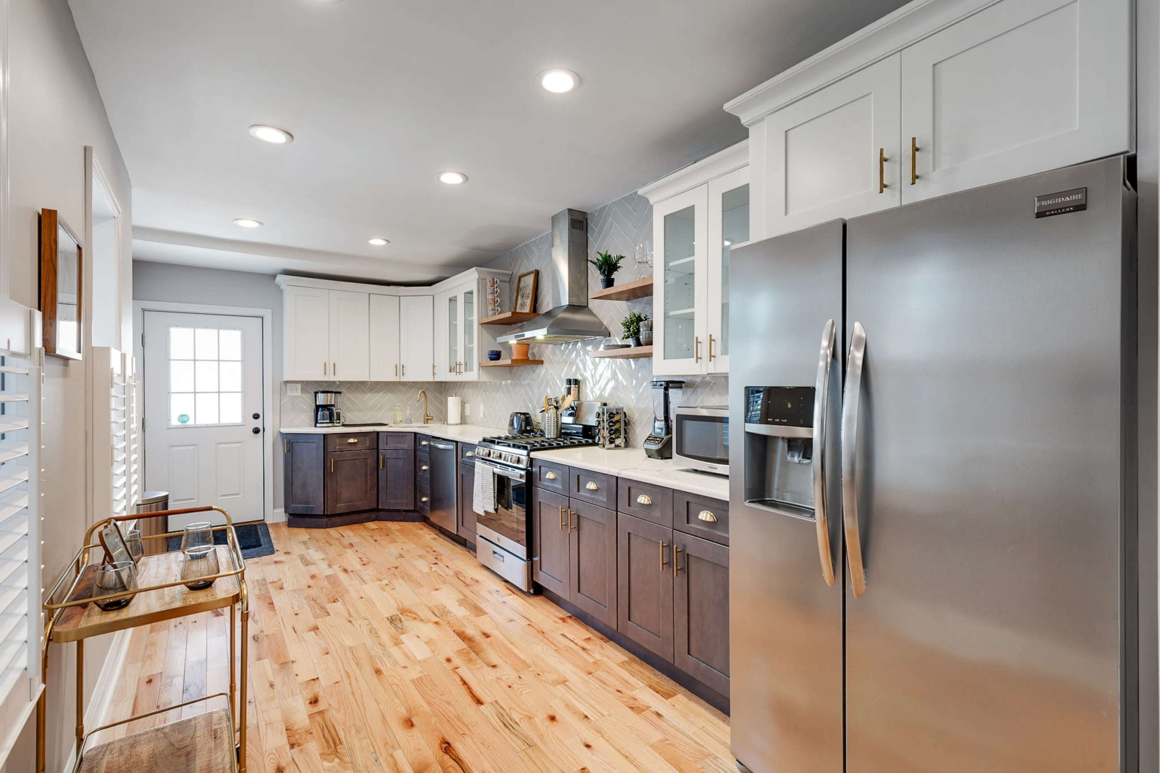 The image shows a modern kitchen featuring a stainless steel refrigerator, a gas stove, and a mix of dark and light cabinetry, with wooden flooring and a door leading outside.