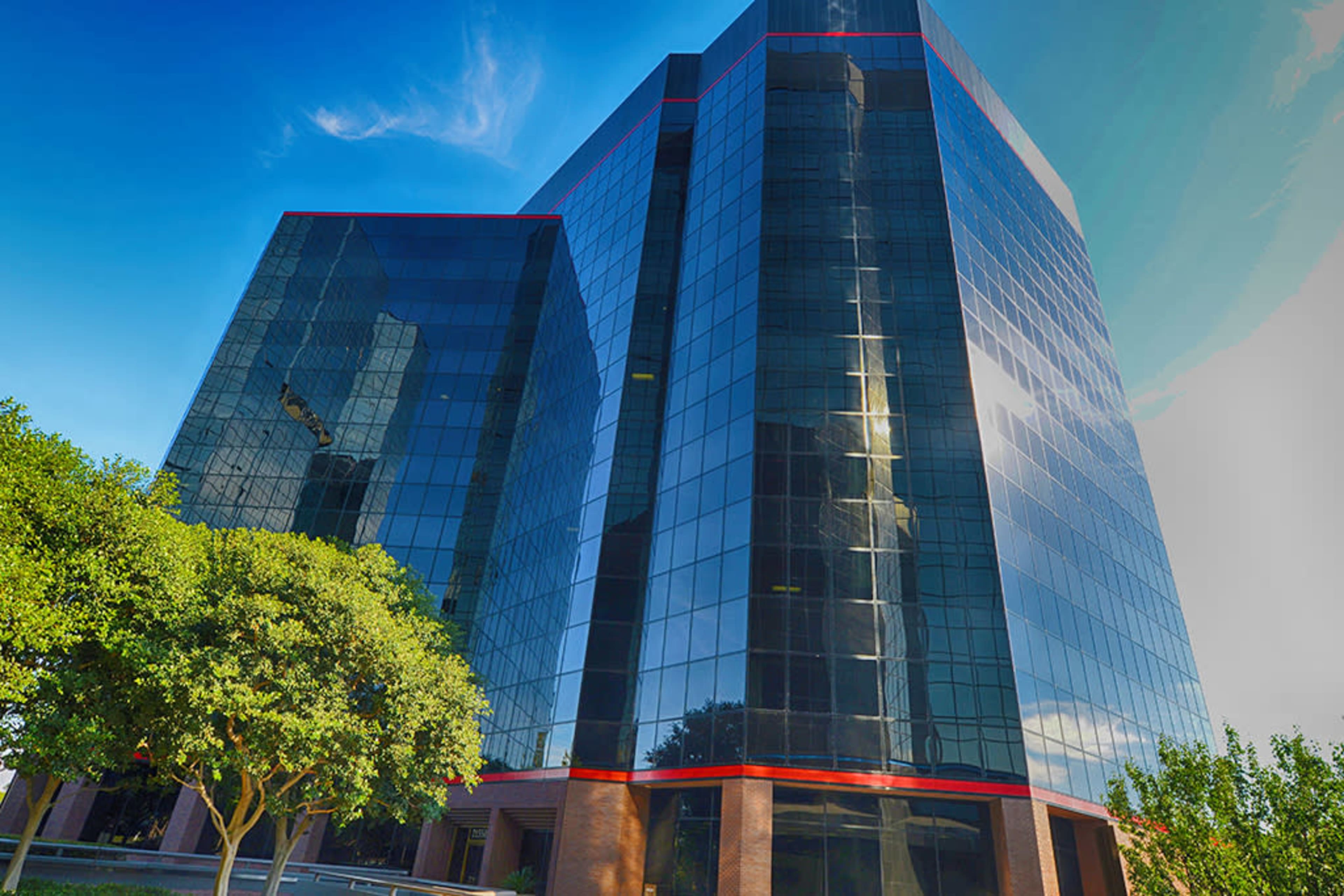 A modern glass office building stands against a clear blue sky, framed by green trees in the foreground.