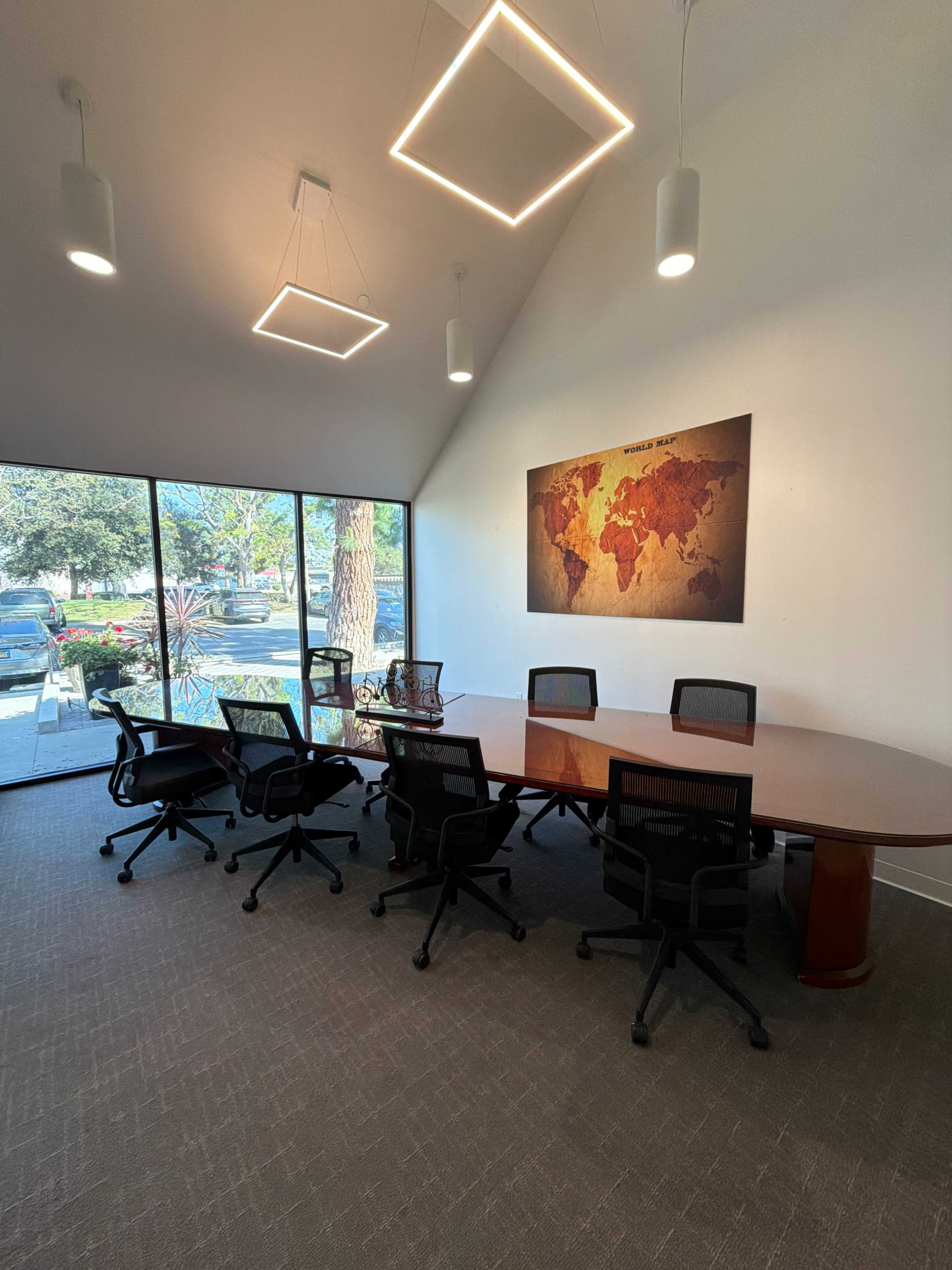 The image shows a modern conference room with a large wooden table surrounded by office chairs, featuring a world map on the wall and large windows letting in natural light.
