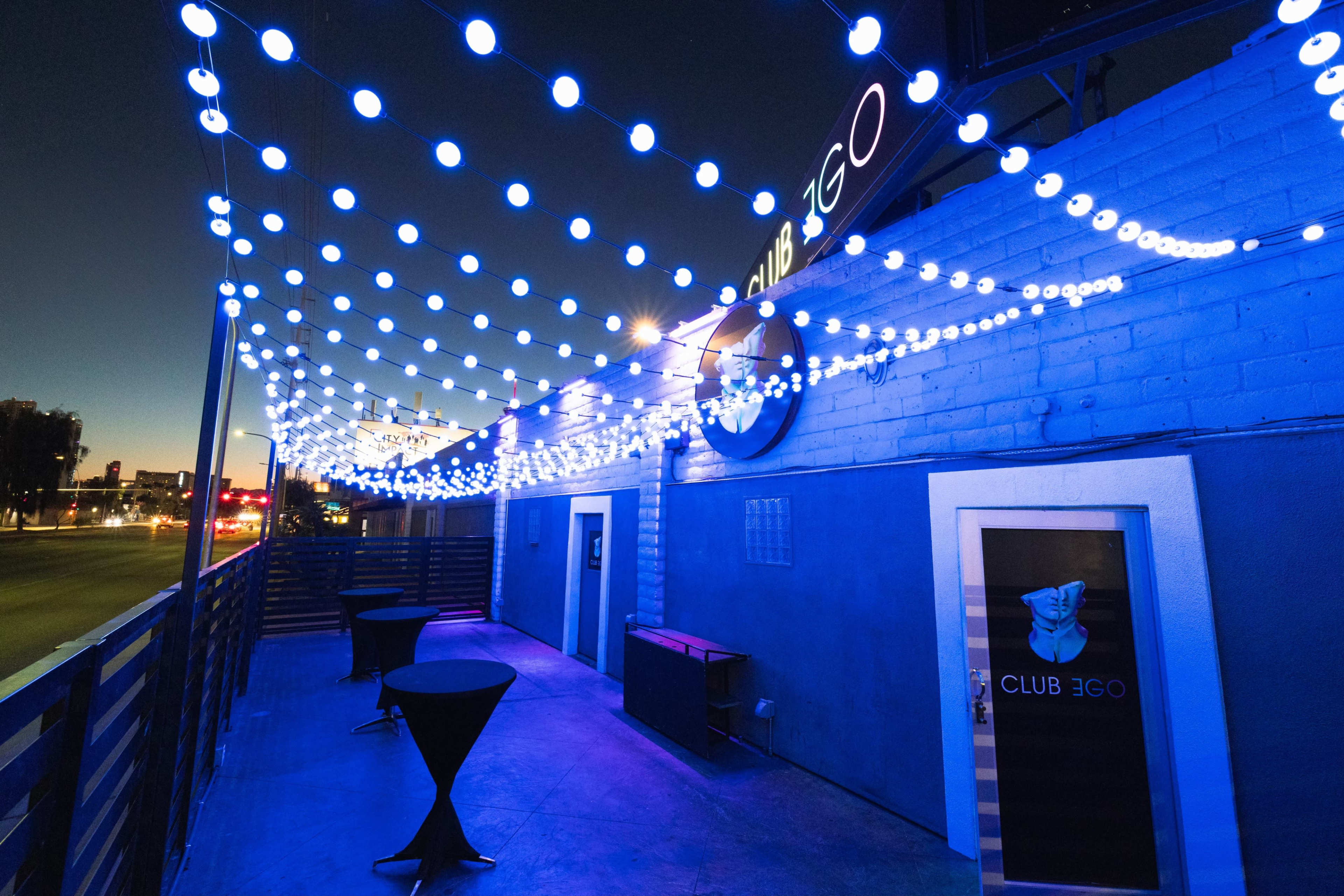 The image shows an outdoor area of a venue named "Club 3GO" adorned with blue string lights and two tall tables.