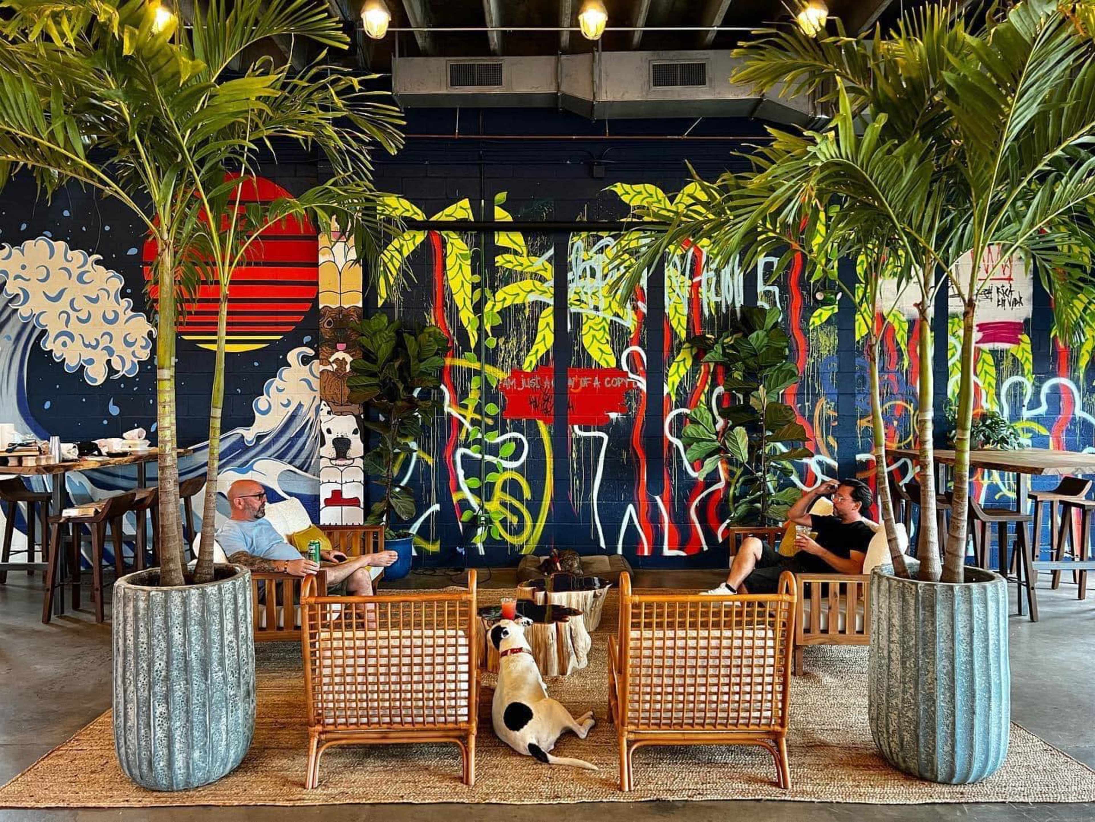 Two people sit in a modern café surrounded by large potted palm trees and a colorful mural on the wall, with a dog resting on the floor between them.