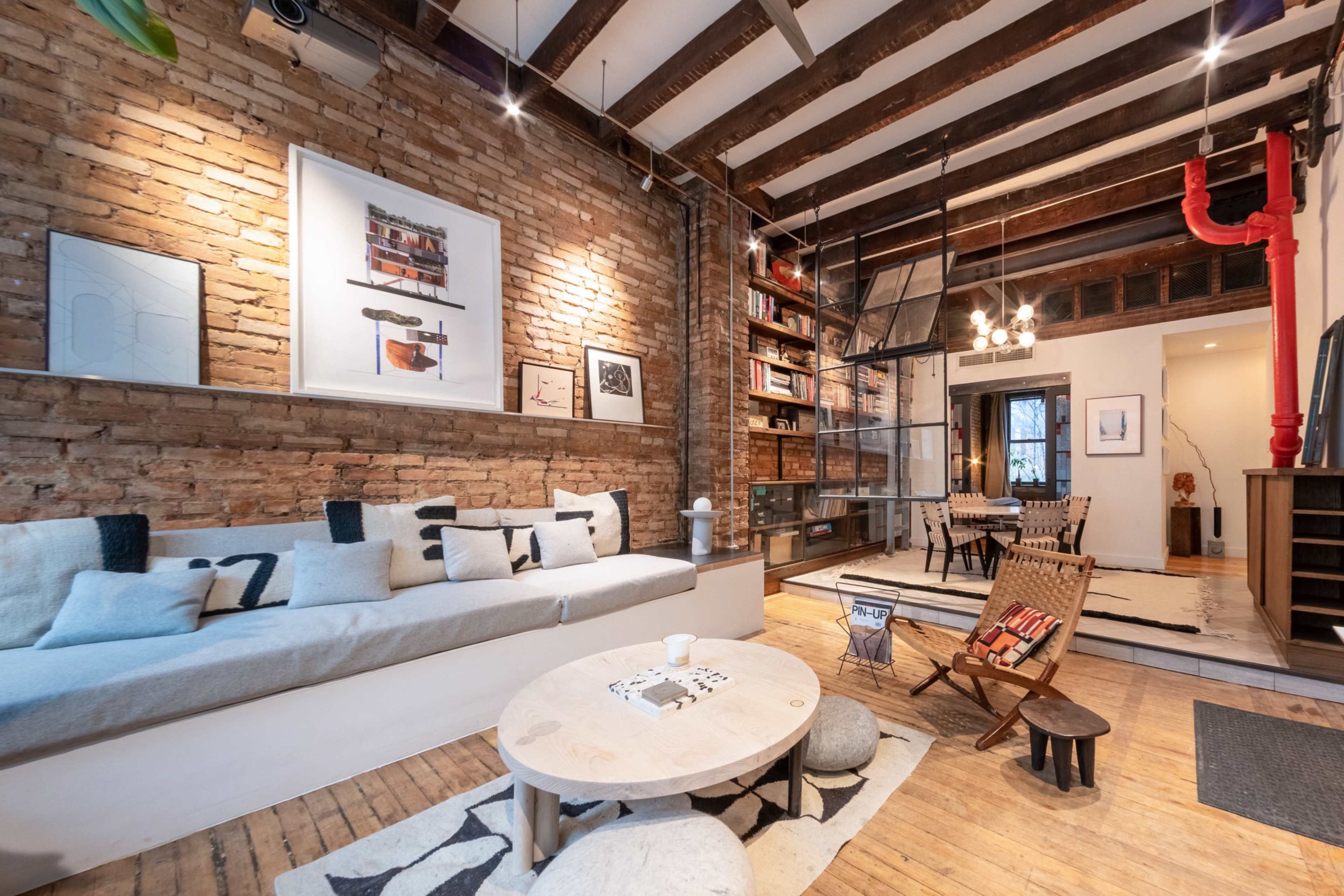 A stylish living room with exposed brick walls, wooden beams, and a mix of modern furniture, including a long couch, a round coffee table, and a wooden chair.
