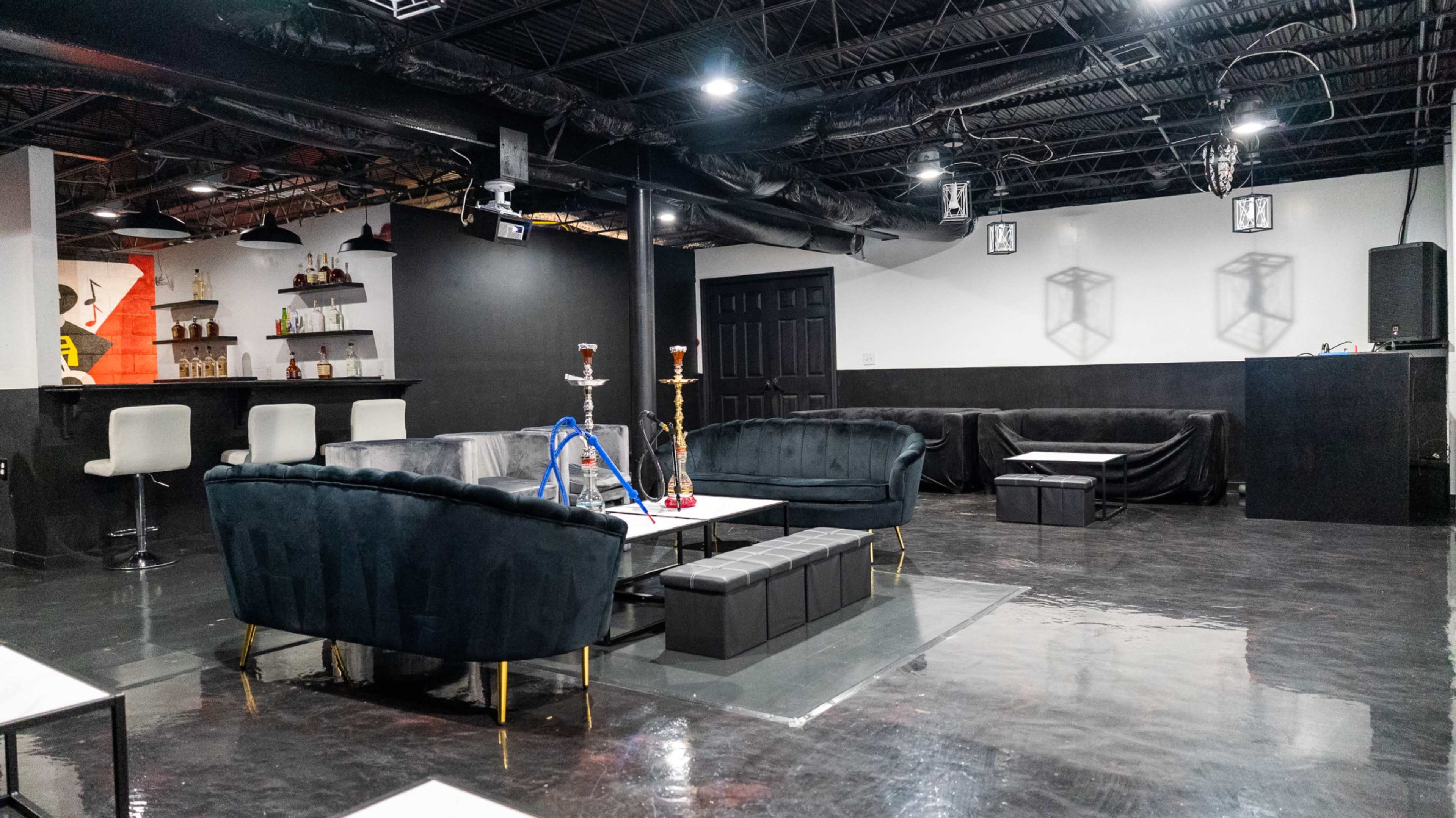 A modern lounge area with dark furnishings, a bar in the background, and hookahs on coffee tables.