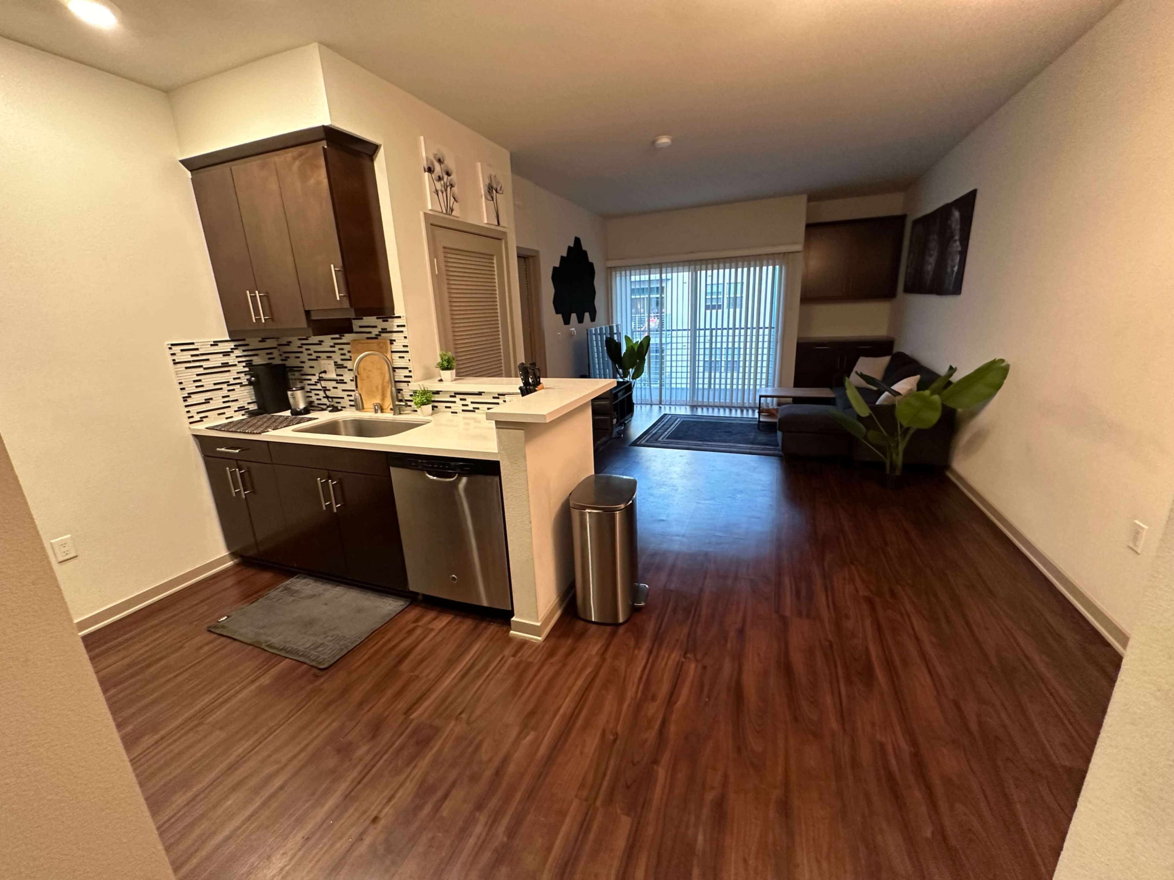 A modern kitchen area with dark cabinets and a small dining space leads into a living area featuring a couch and large window with blinds.