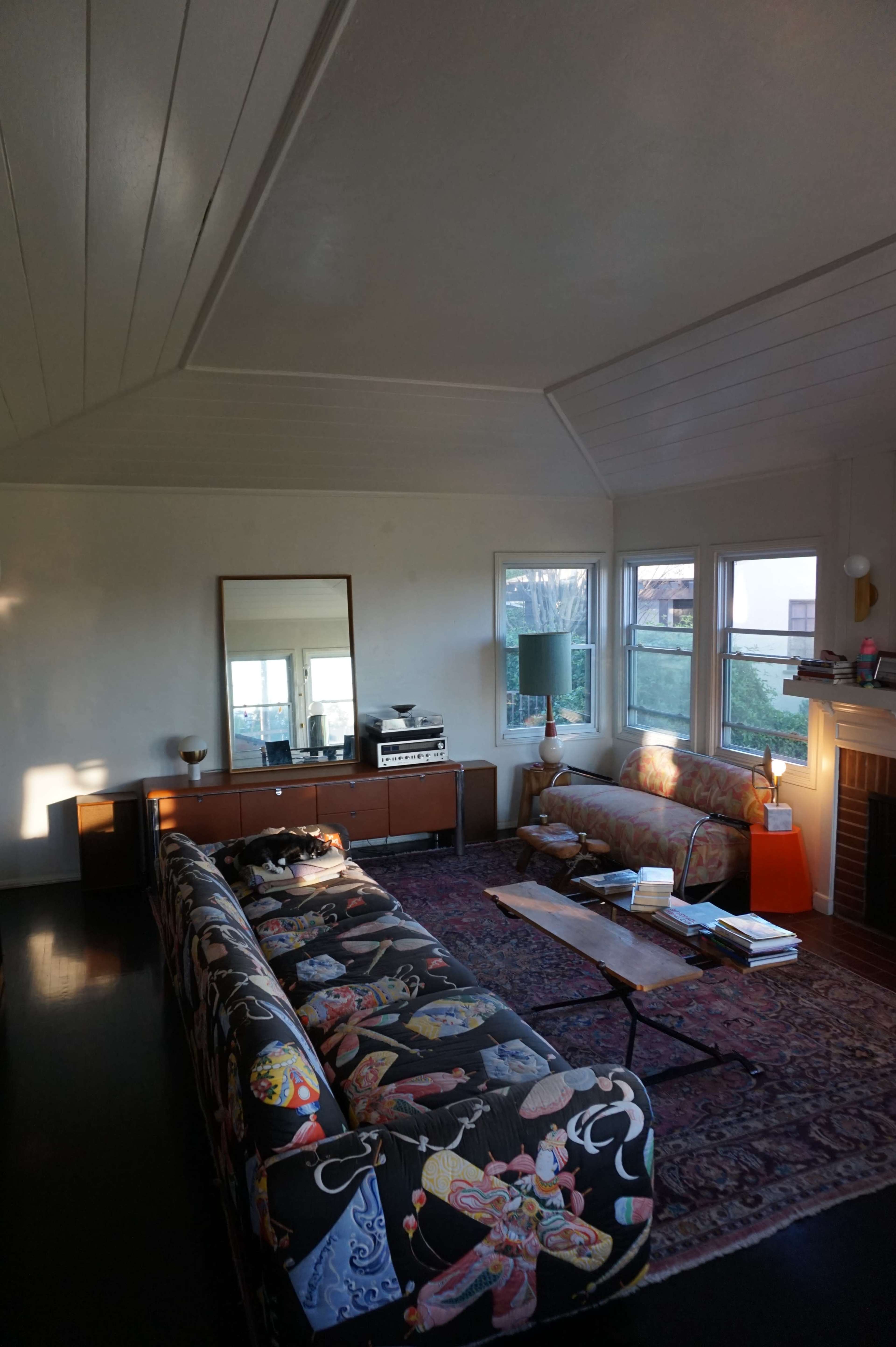 The room features a patterned sofa and a wooden coffee table, with large windows allowing natural light and a view of the outdoors.