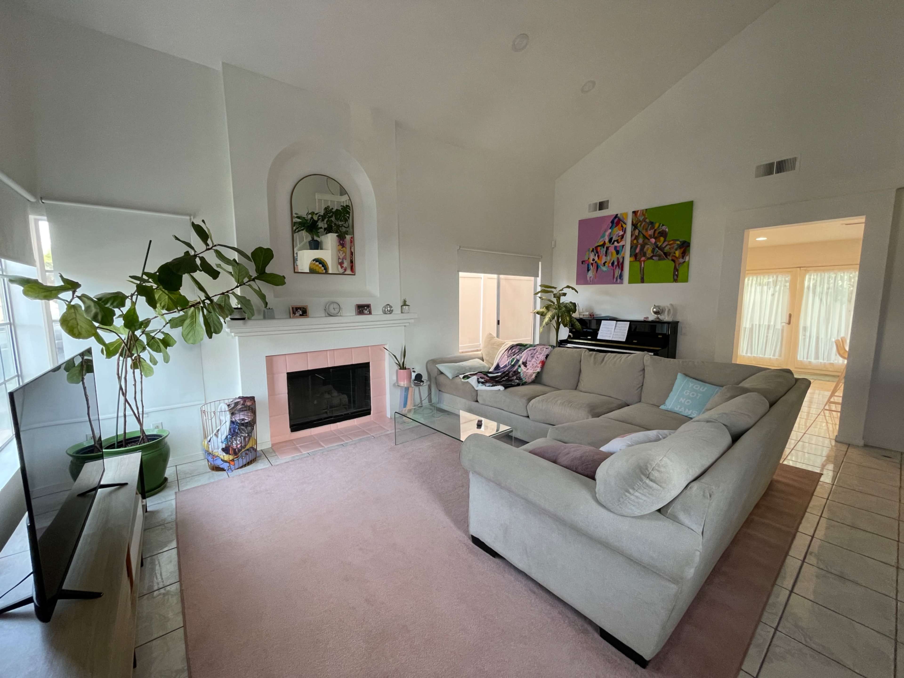 The living room features a large beige sectional sofa, a fireplace with a pink surround, and large windows allowing natural light to fill the space.