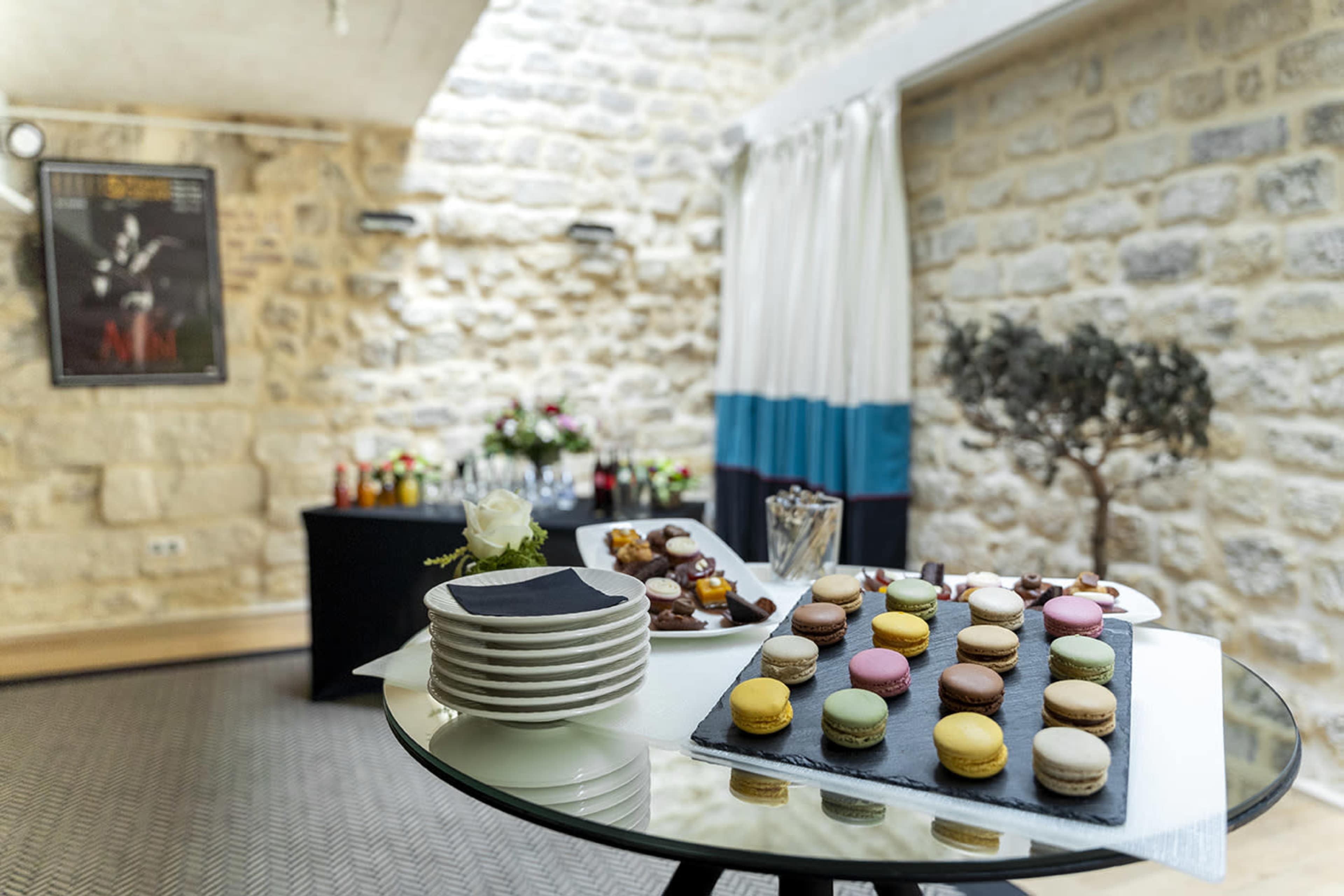 A table set with plates and colorful macarons, alongside a buffet featuring drinks and floral arrangements in a stone-walled room.