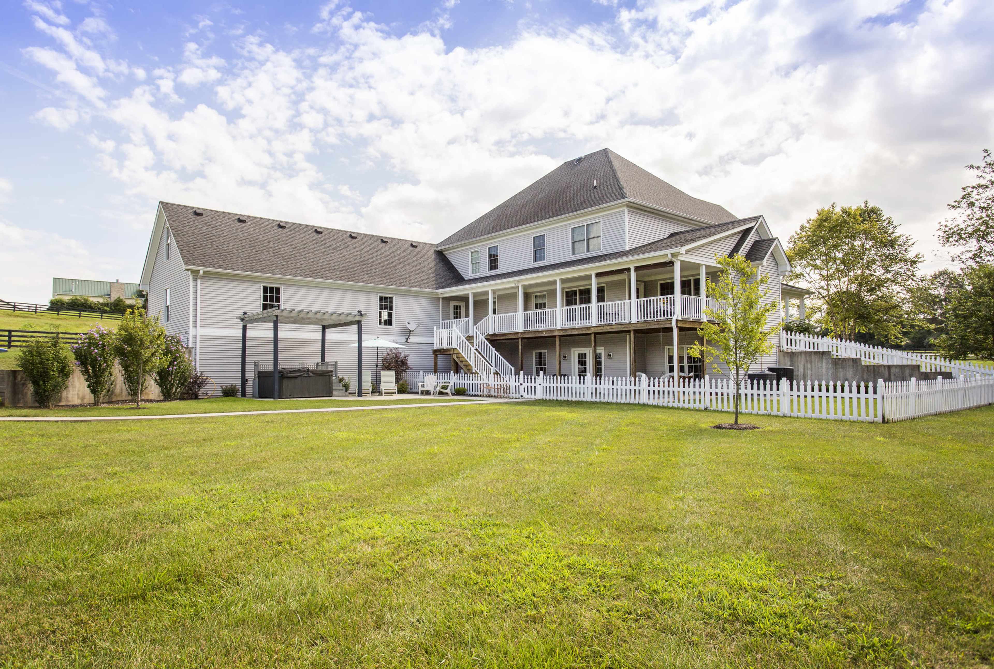 A large two-story house with a peaked roof and a wraparound porch is set on a grassy lawn surrounded by a white picket fence.