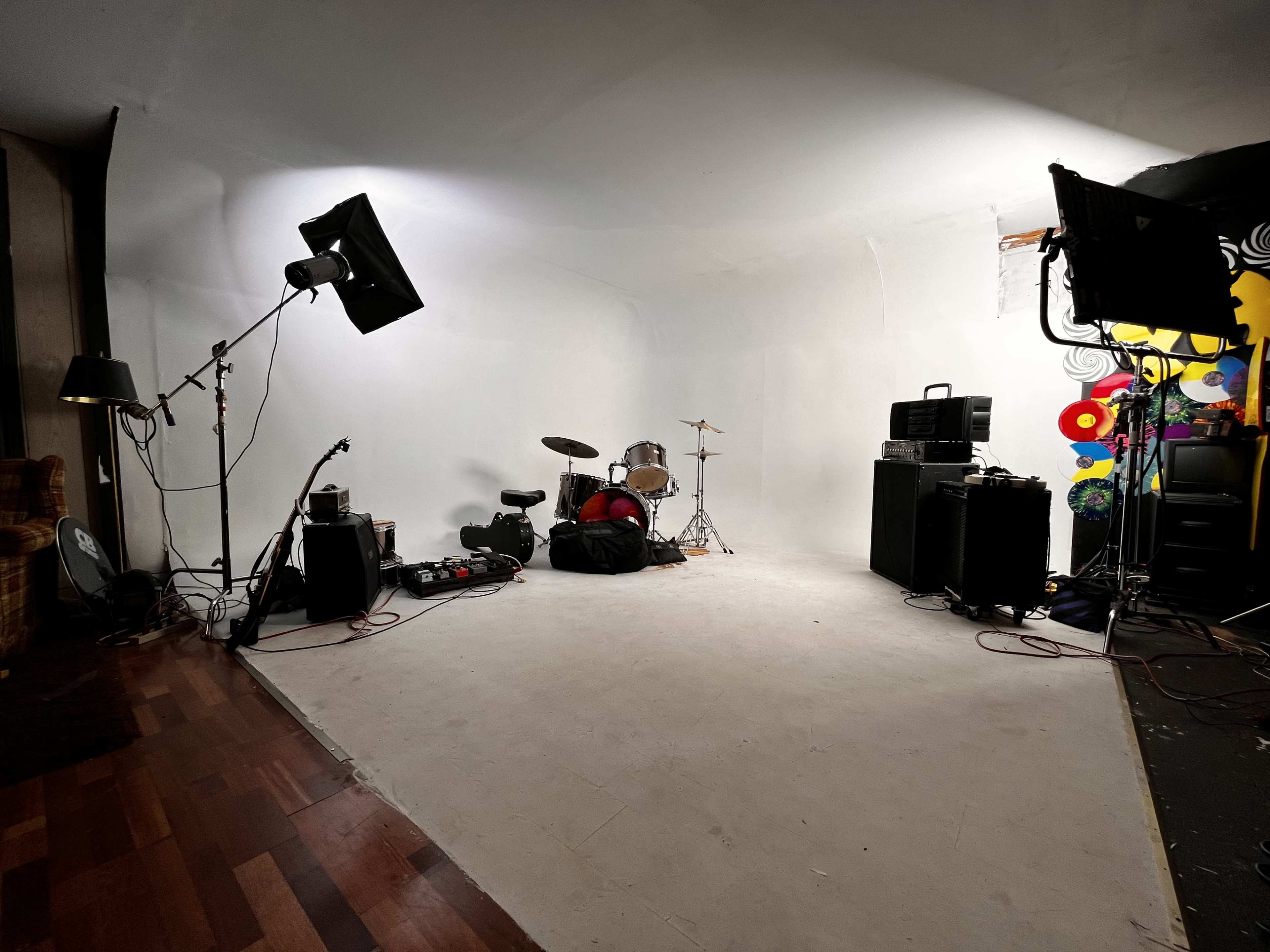 An empty music rehearsal space featuring a drum set, amplifiers, a guitar on a stand, and studio lighting against a plain background.